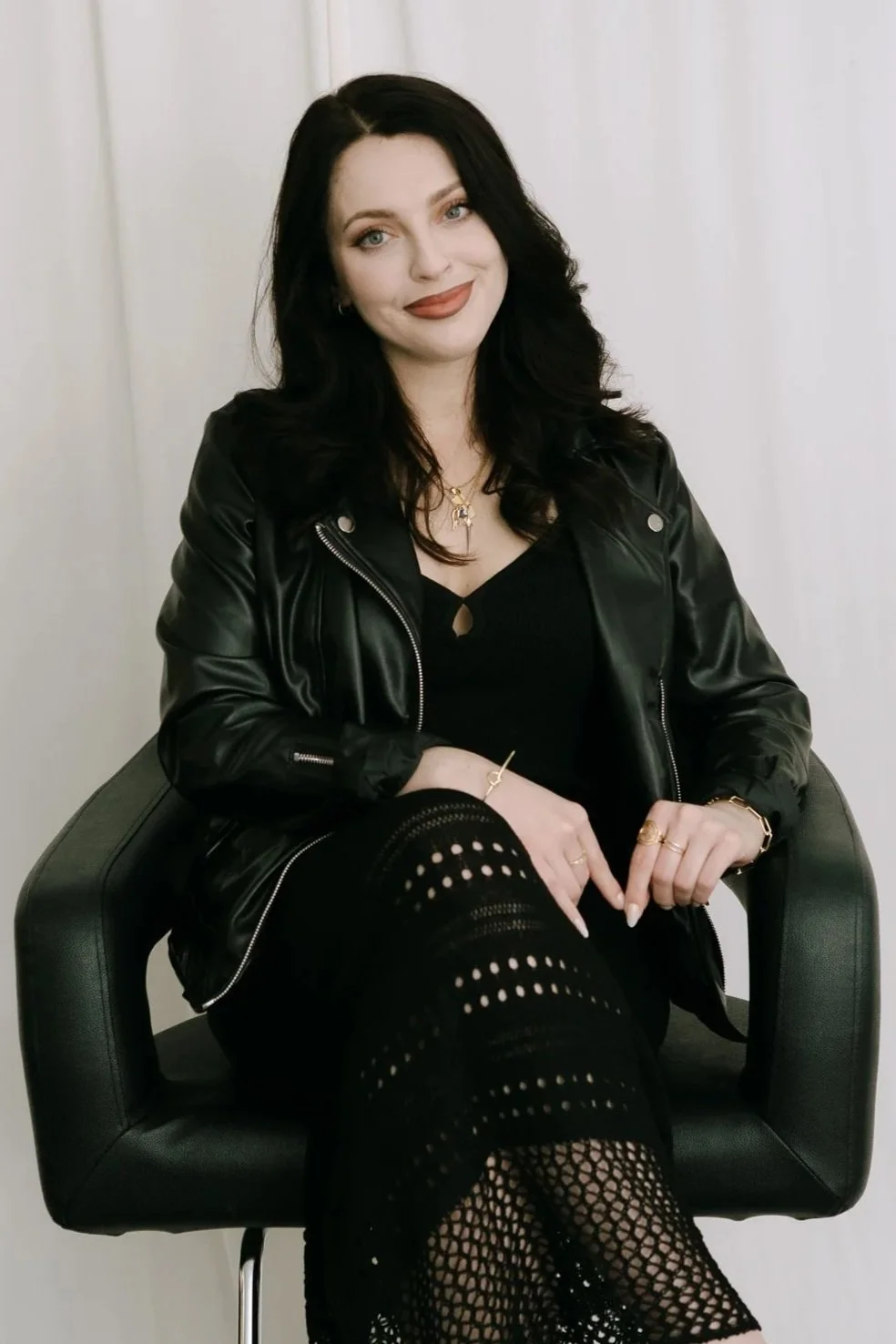 A woman with dark hair, wearing a black leather jacket, black dress, and fishnet tights, sitting on a black chair against a plain light-colored background.