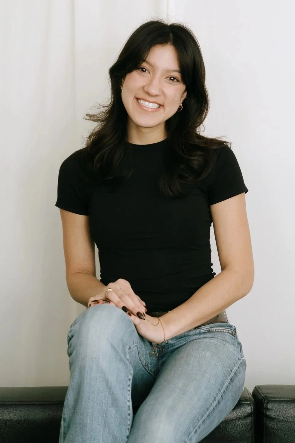 Smiling woman with dark hair, wearing a black t-shirt and jeans, sitting on a black couch with a white background.