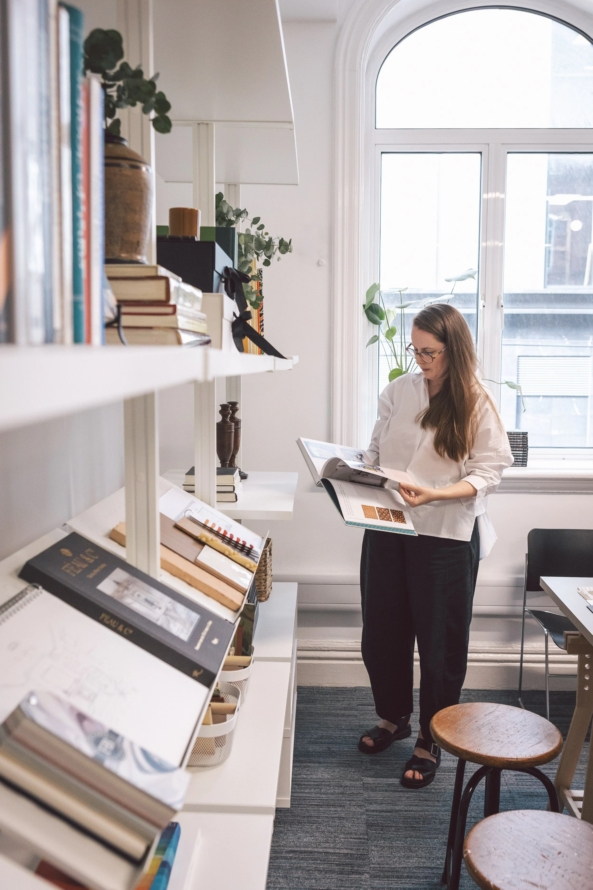A woman with long brown hair, glasses, and wearing a white blouse and black pants, standing in a brightly lit room with large arched window, browsing through a magazine. The room has white shelves filled with books and decorative items, and few wooden stools near a white desk.