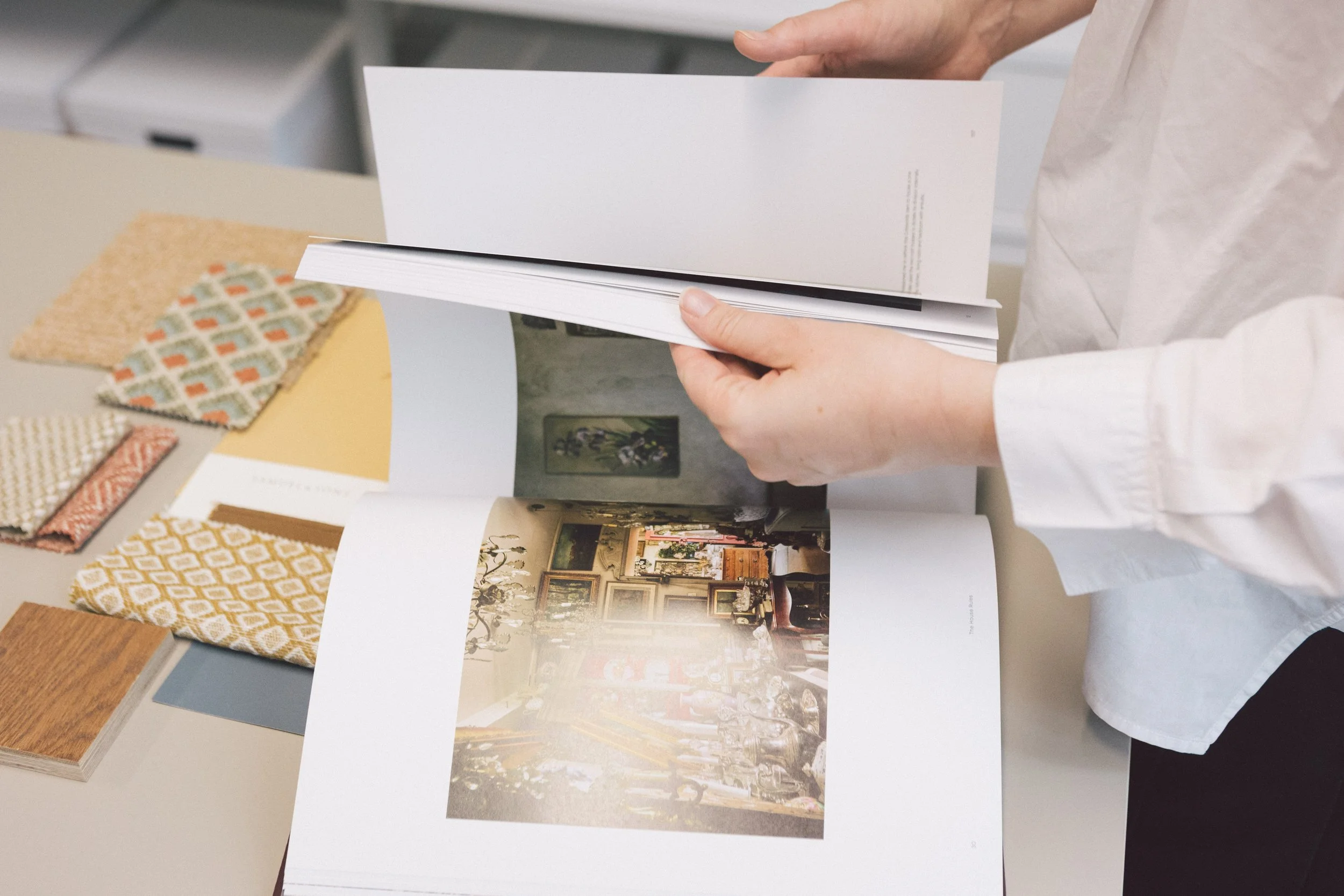 Person inspecting interior design or furniture catalog on table, surrounded by fabric samples and books.