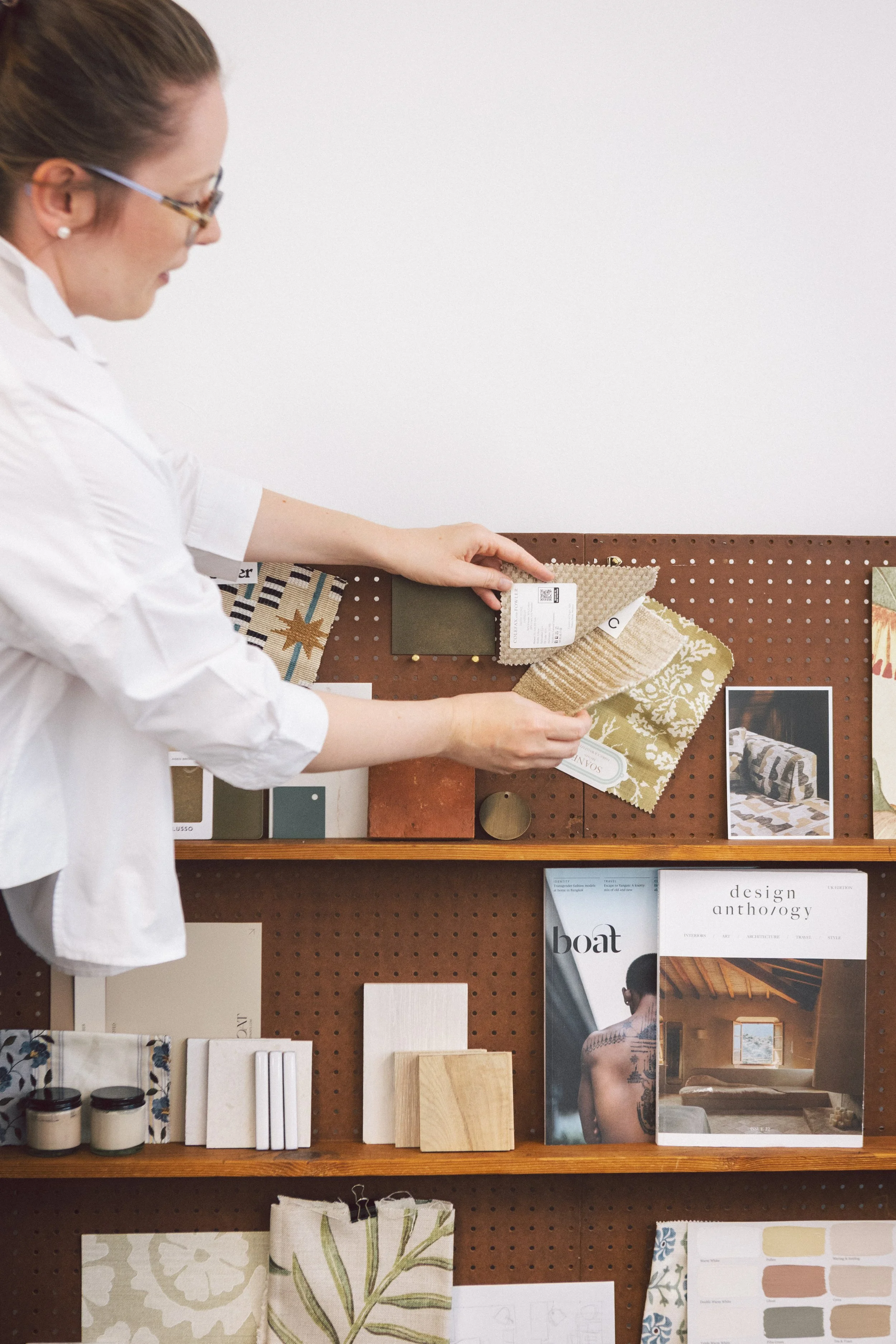 A woman wearing glasses and a white shirt looking at fabric swatches on a pegboard wall display, with magazines and fabric samples on a wooden table below.