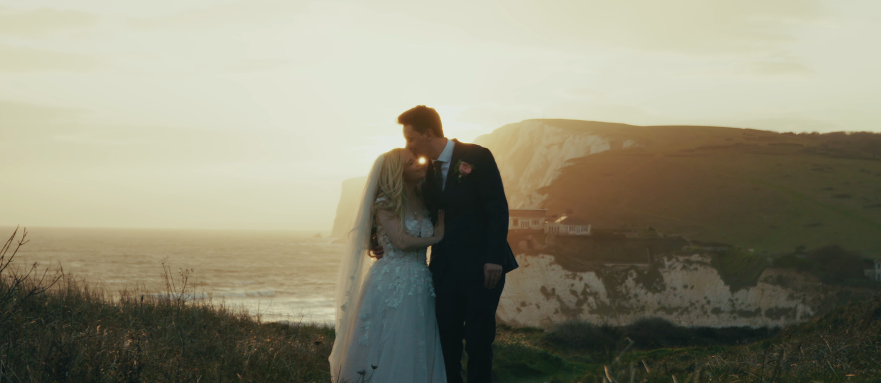 A bride and groom embracing on a grassy cliffside at sunset with ocean waves and white cliffs in the background.