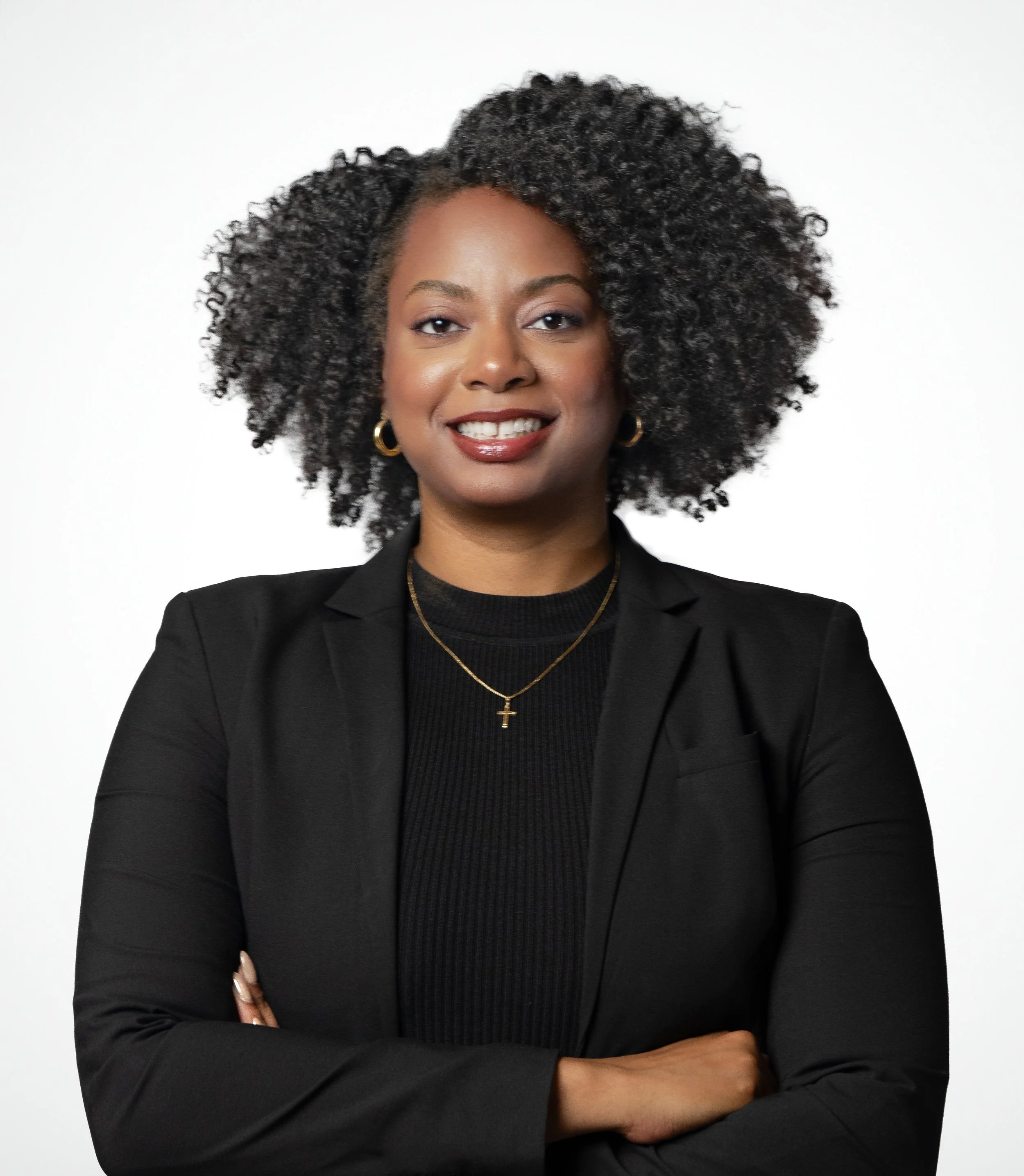 A professional woman with curly hair, wearing a black blazer, black top, gold jewelry, and smiling with arms crossed.