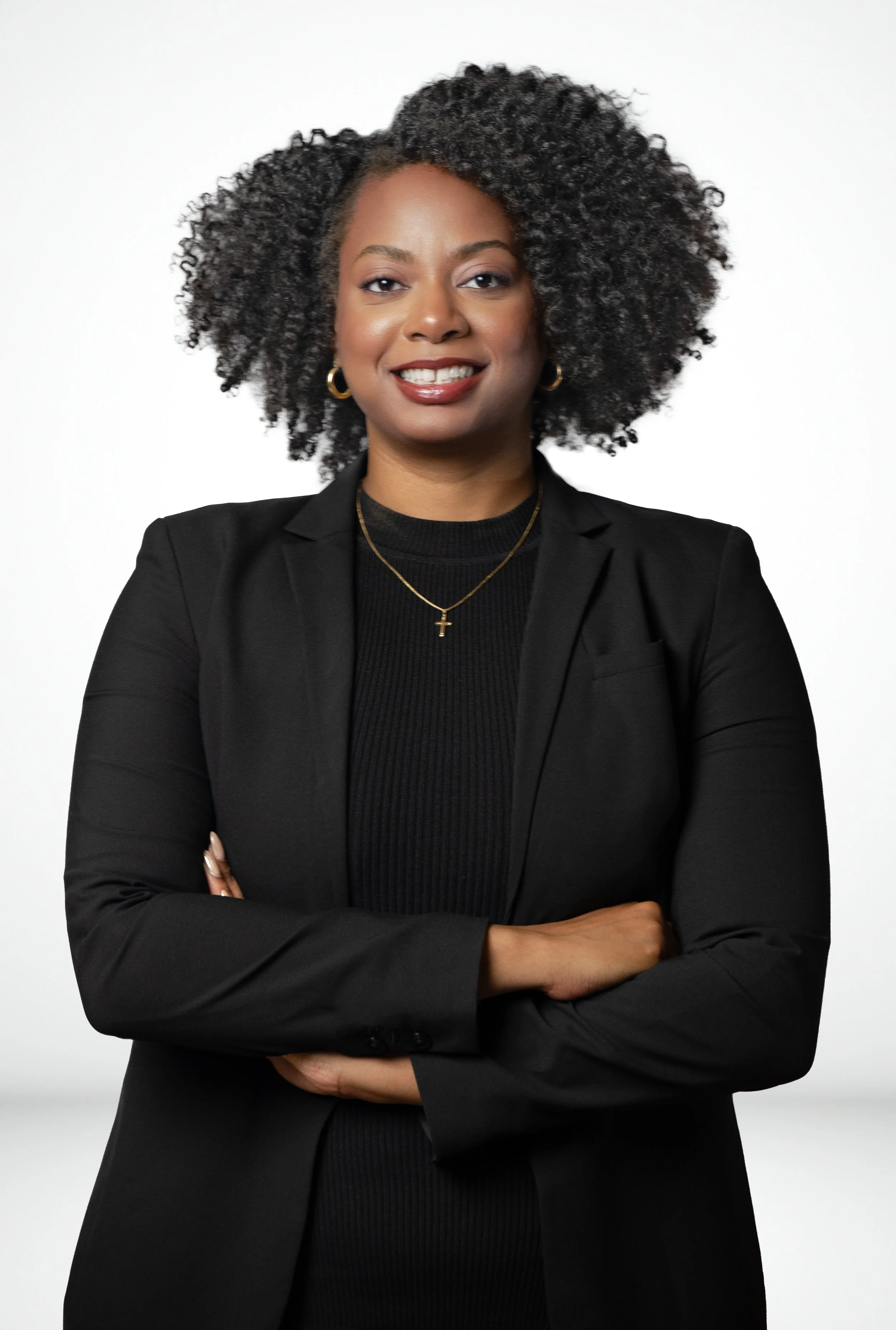 A confident African American woman with curly hair, wearing a black blazer and black top, standing with arms crossed against a plain white background.