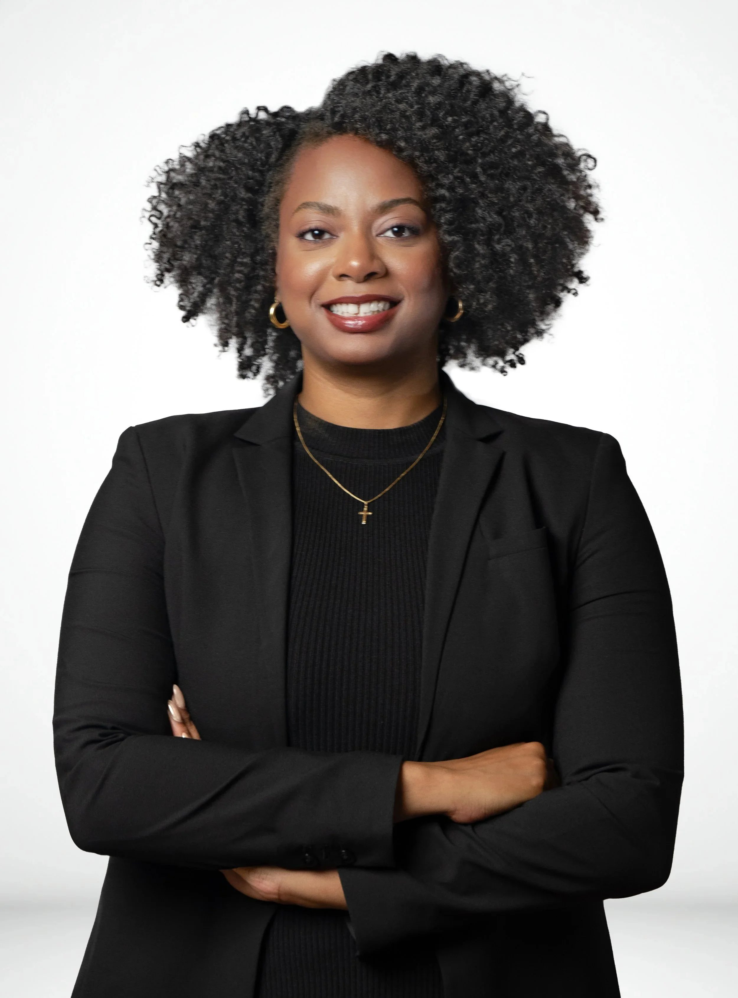 Portrait of a confident African American woman with curly hair, wearing a black blazer and jewelry, standing with arms crossed against a white background.