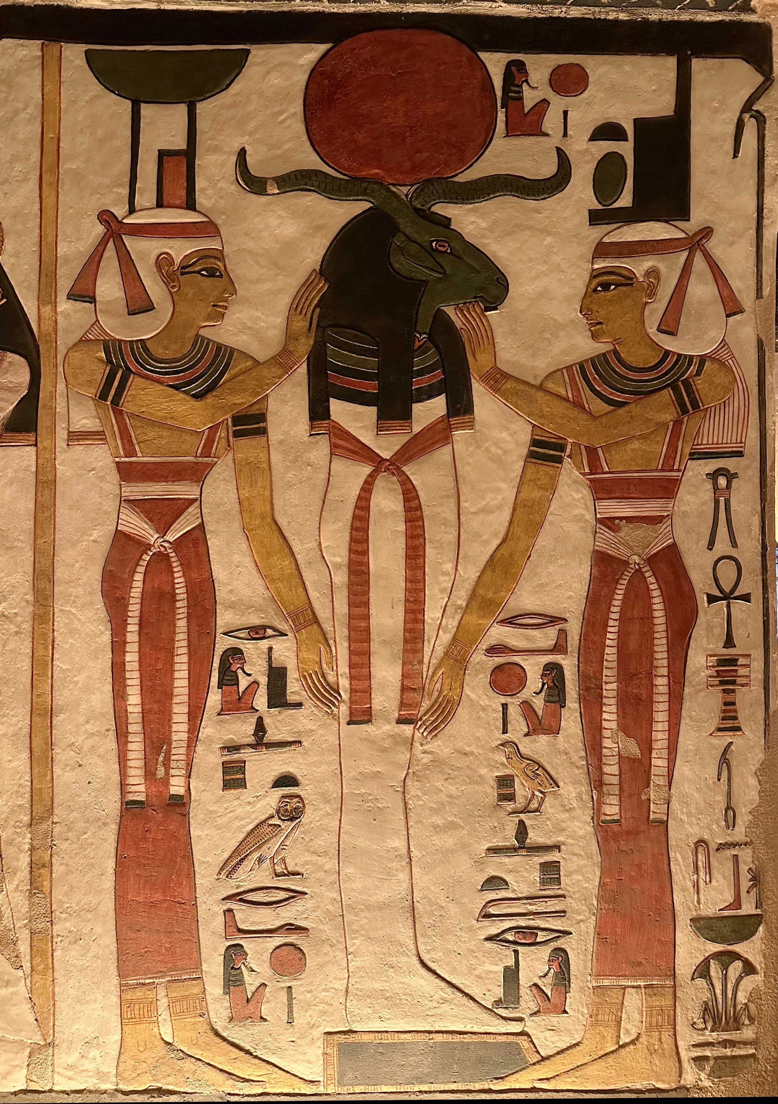 Painted ancient Egyptian relief of a ram-headed mummy with solar disk between two goddesses.