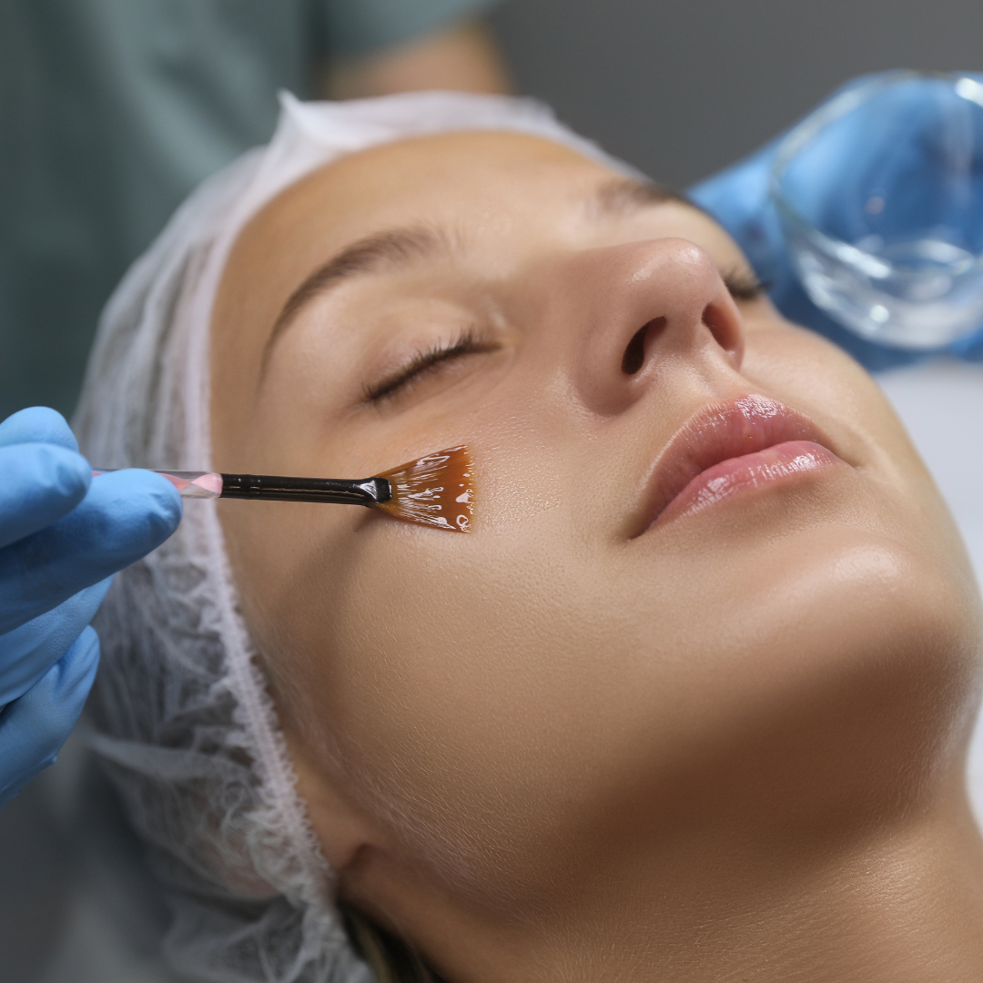 A woman receives a facial treatment with a skincare product applied using a brush by a esthetician, lying with eyes closed.