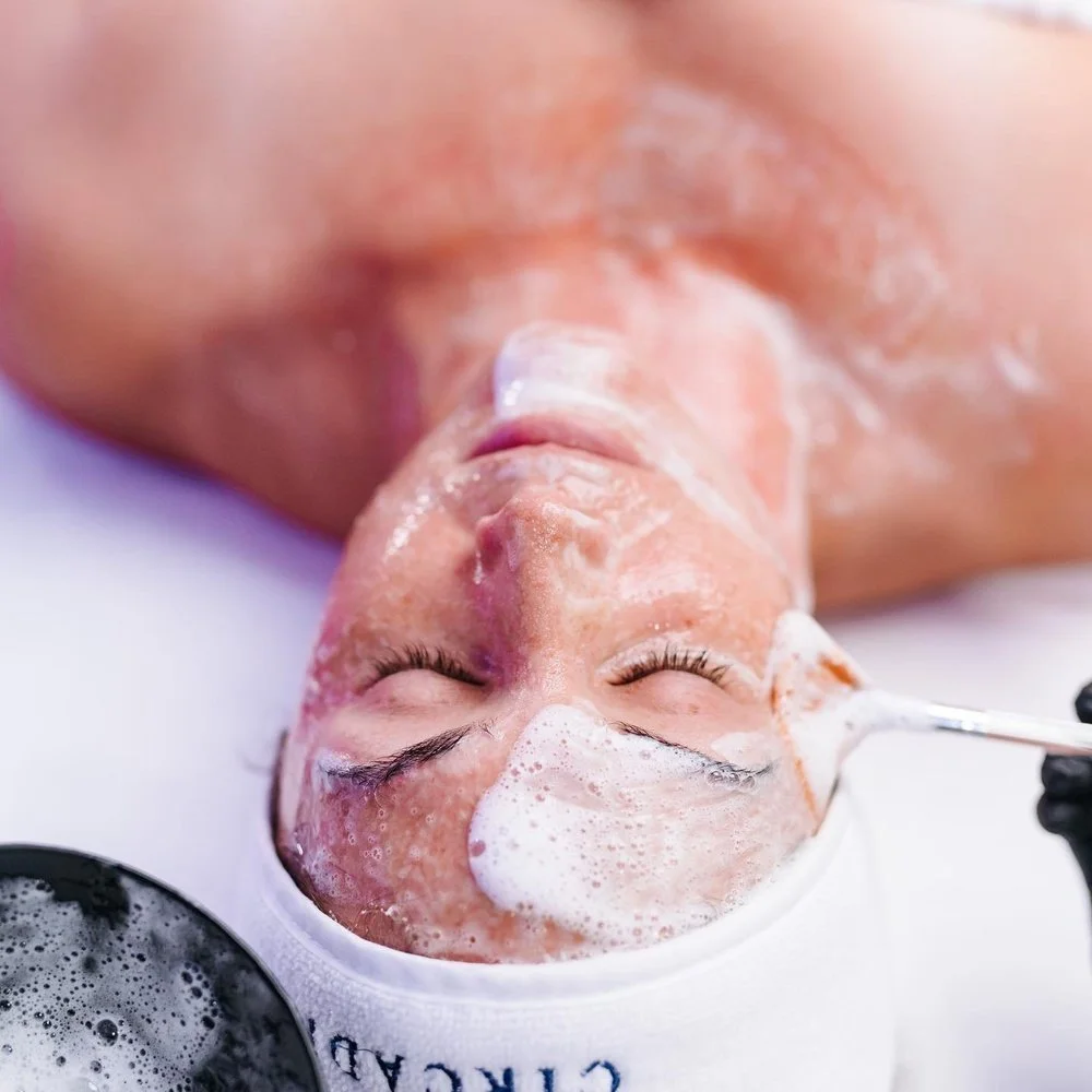 A person receiving a facial treatment, with foam and bubbles on their face, lying down with eyes closed.