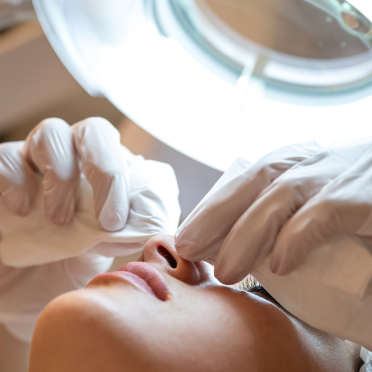 Person receiving a cosmetic or dermatological treatment, with a healthcare professional wearing gloves near the person's nose and face, under a bright examination light.