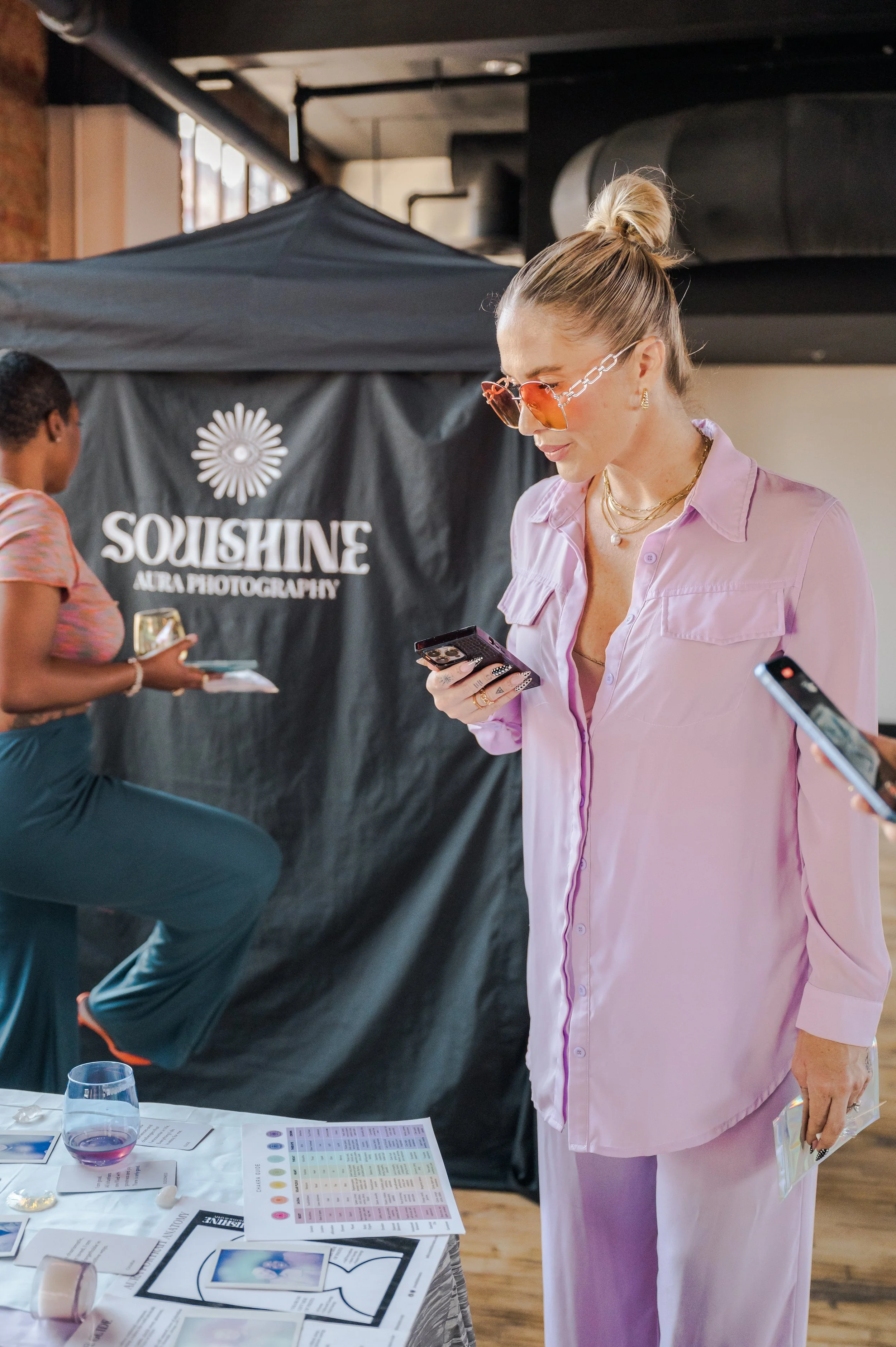 A woman with long blonde hair in a green sleeveless top and bright pink pants using a camera or imaging device, focusing on a subject or scene through the viewfinder.