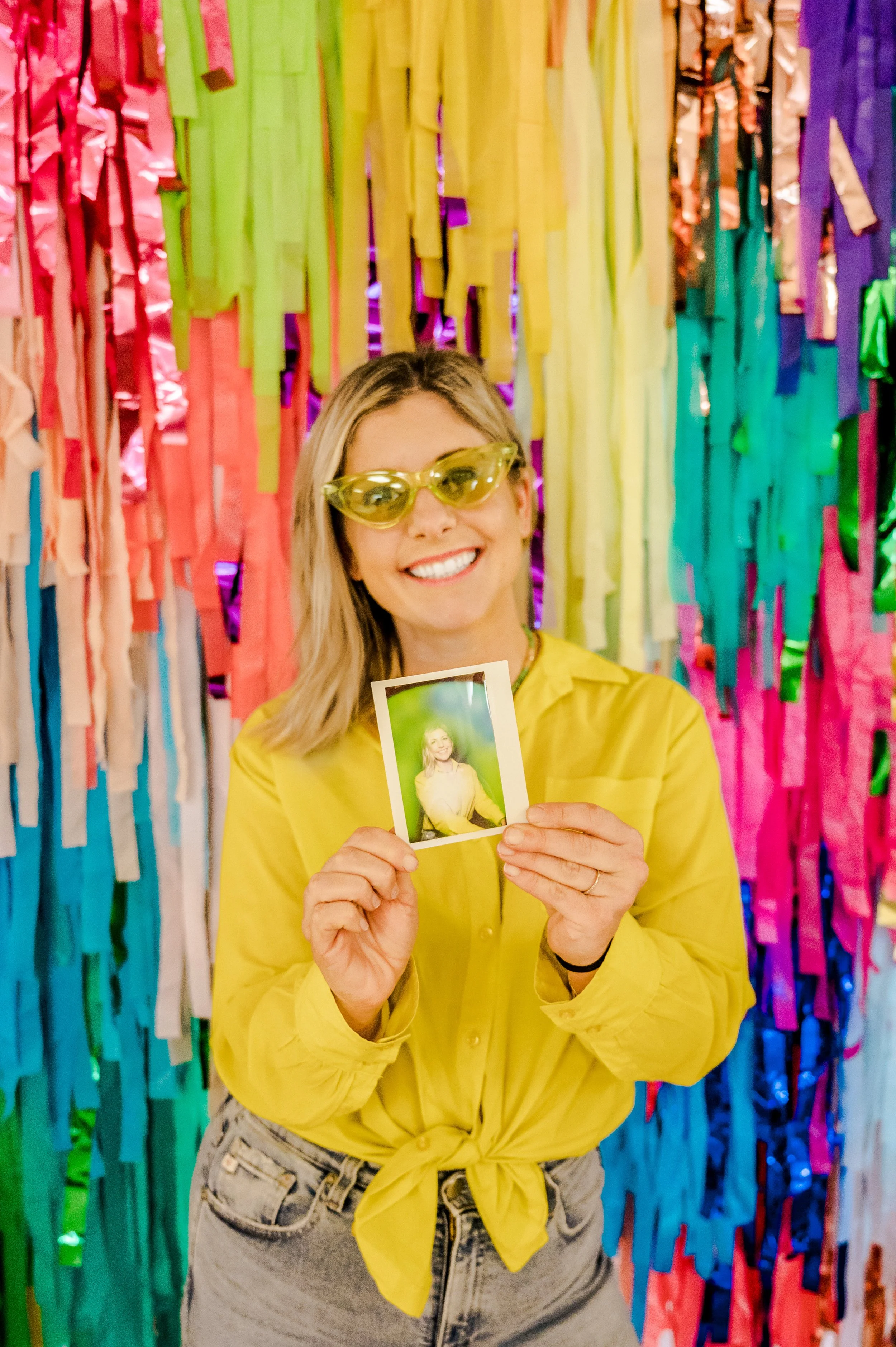 Two women are smiling and holding Polaroid photos, sitting on a pink shopping cart, with colorful flowers in the background.