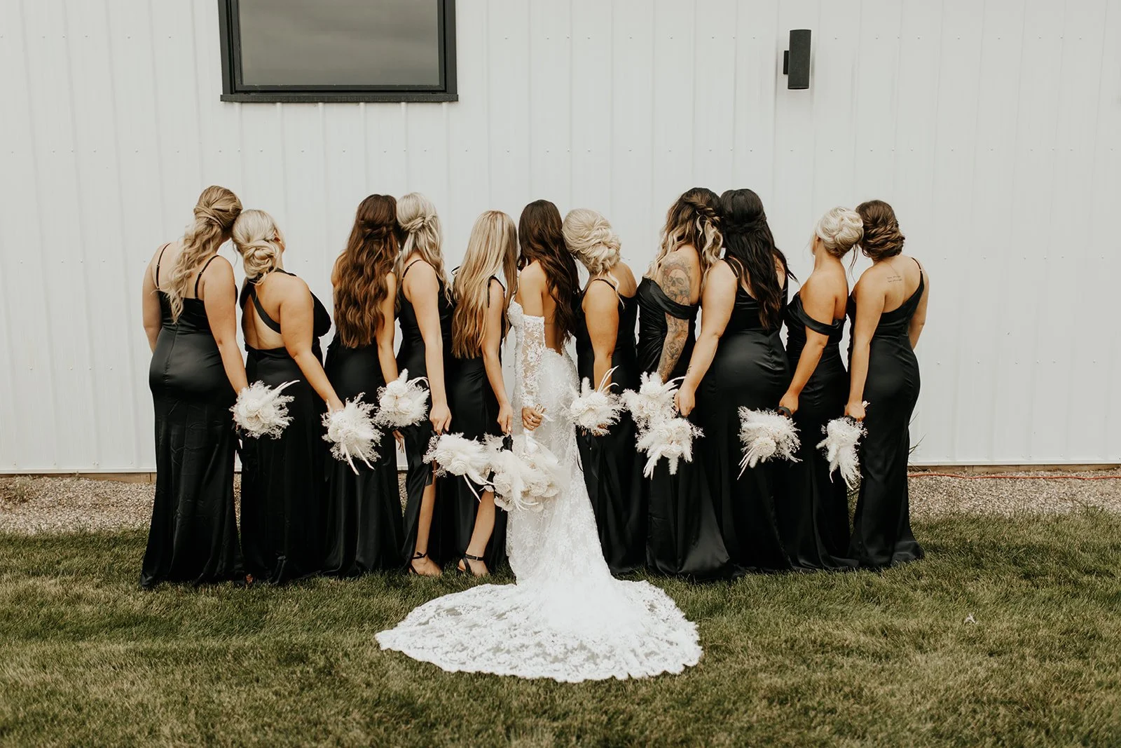A bride in a white wedding dress surrounded by eleven bridesmaids in black dresses, standing outdoors on a grassy area in front of a white wall with a small window.