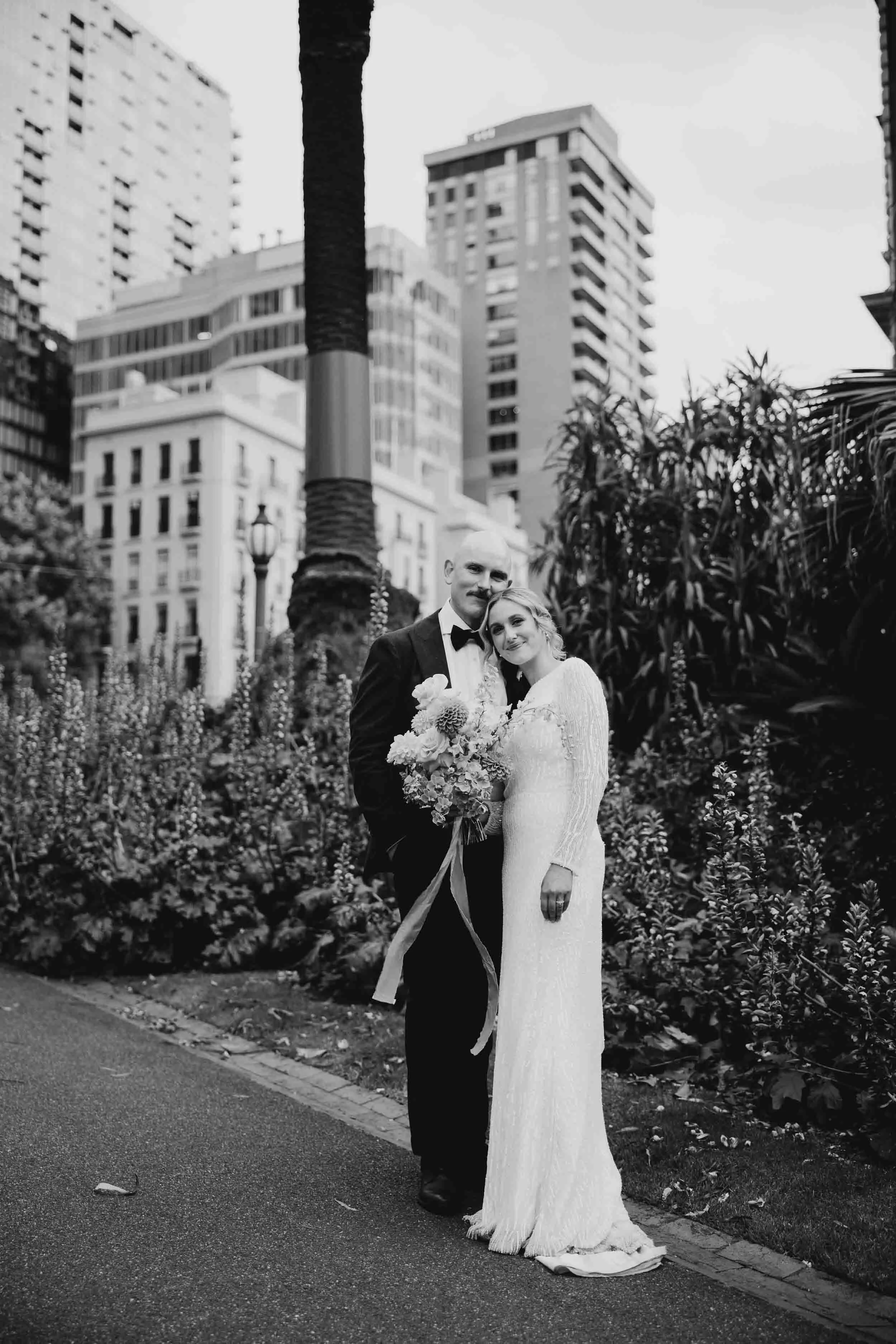 In black and white, Aisling leans into Alex on a garden path at Fitzroy Gardens, the Melbourne skyline rising behind the palms and garden beds.