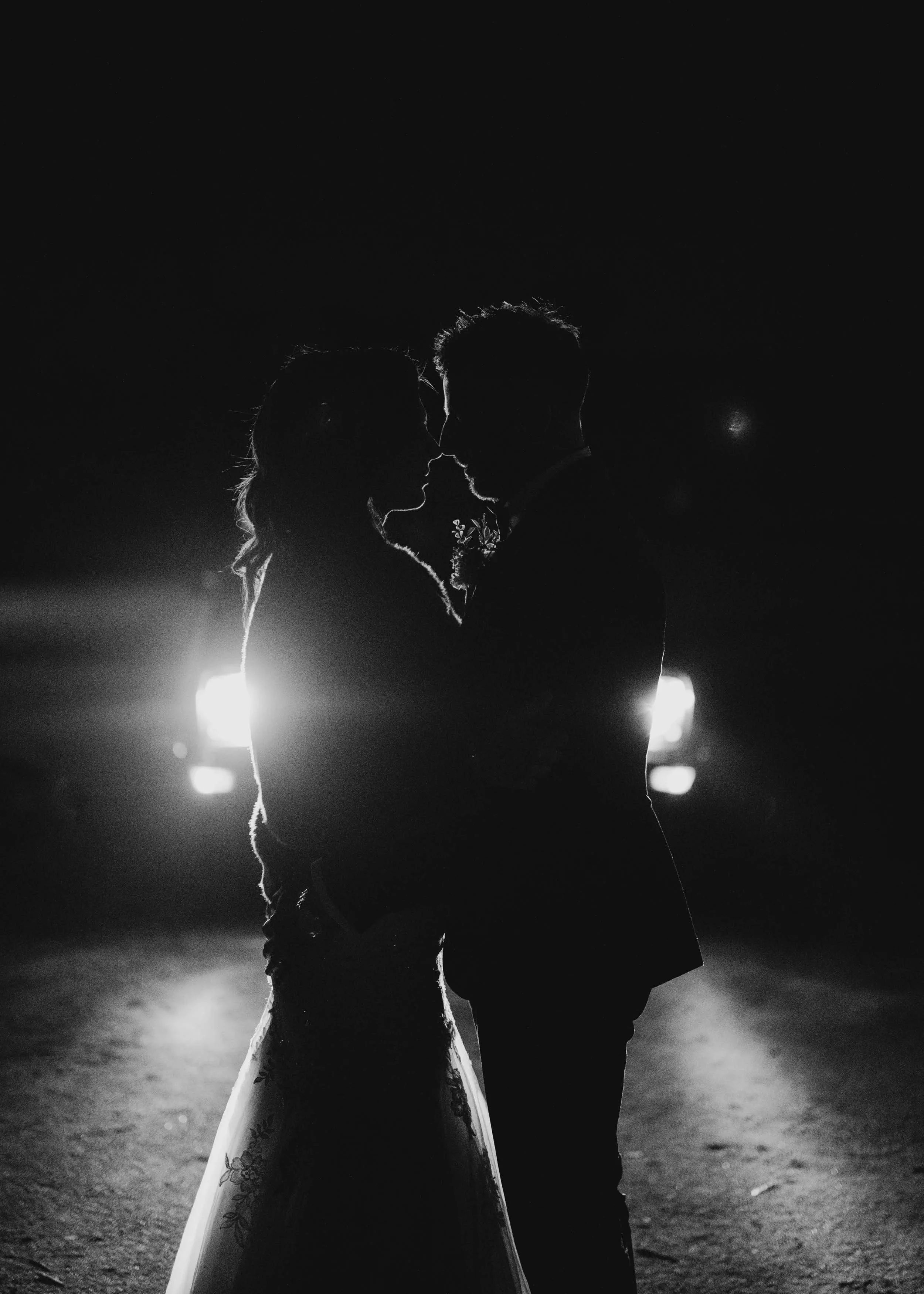 Carmel and Anthony lean into a kiss, rim-lit by the headlights of a Mustang during a thunderstorm at Warrawong Estate.