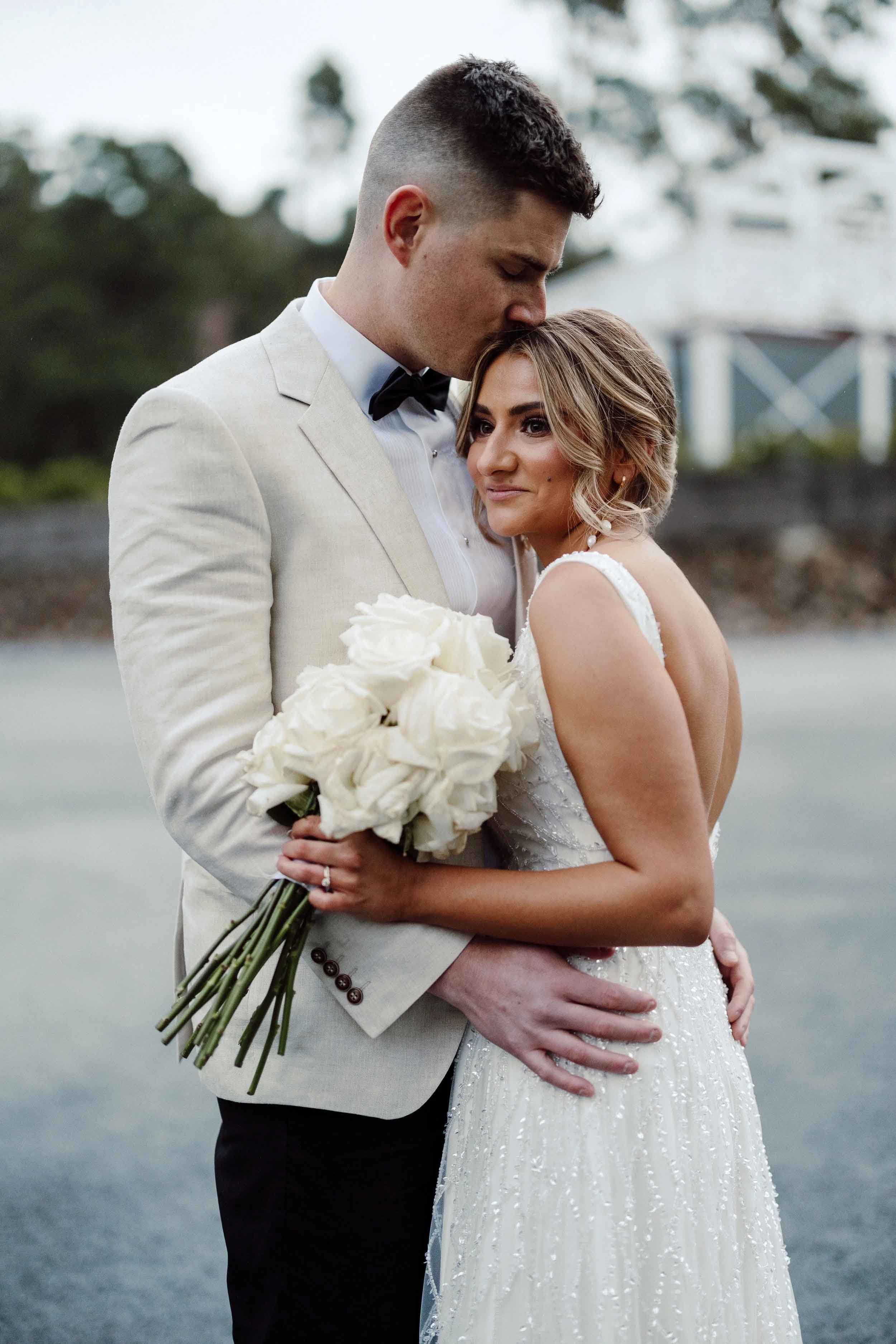 Nick kisses the top of Nicola's head as she leans into him, her white rose bouquet held between them, at Bramleigh Estate.