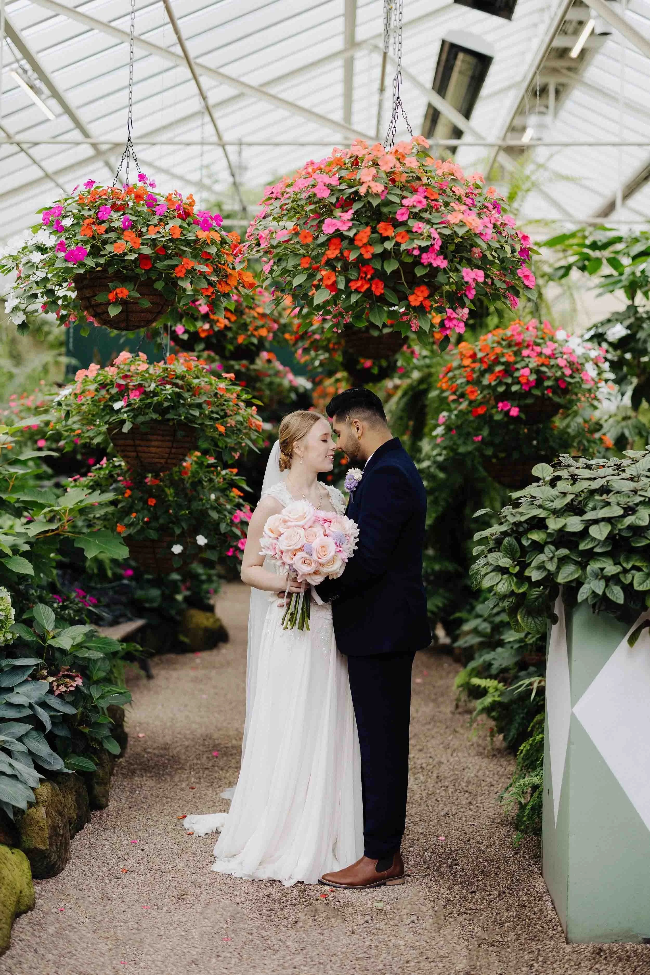 Louise and Sid stand forehead to forehead beneath hanging baskets of pink and orange impatiens inside the Fitzroy Gardens Conservatory.