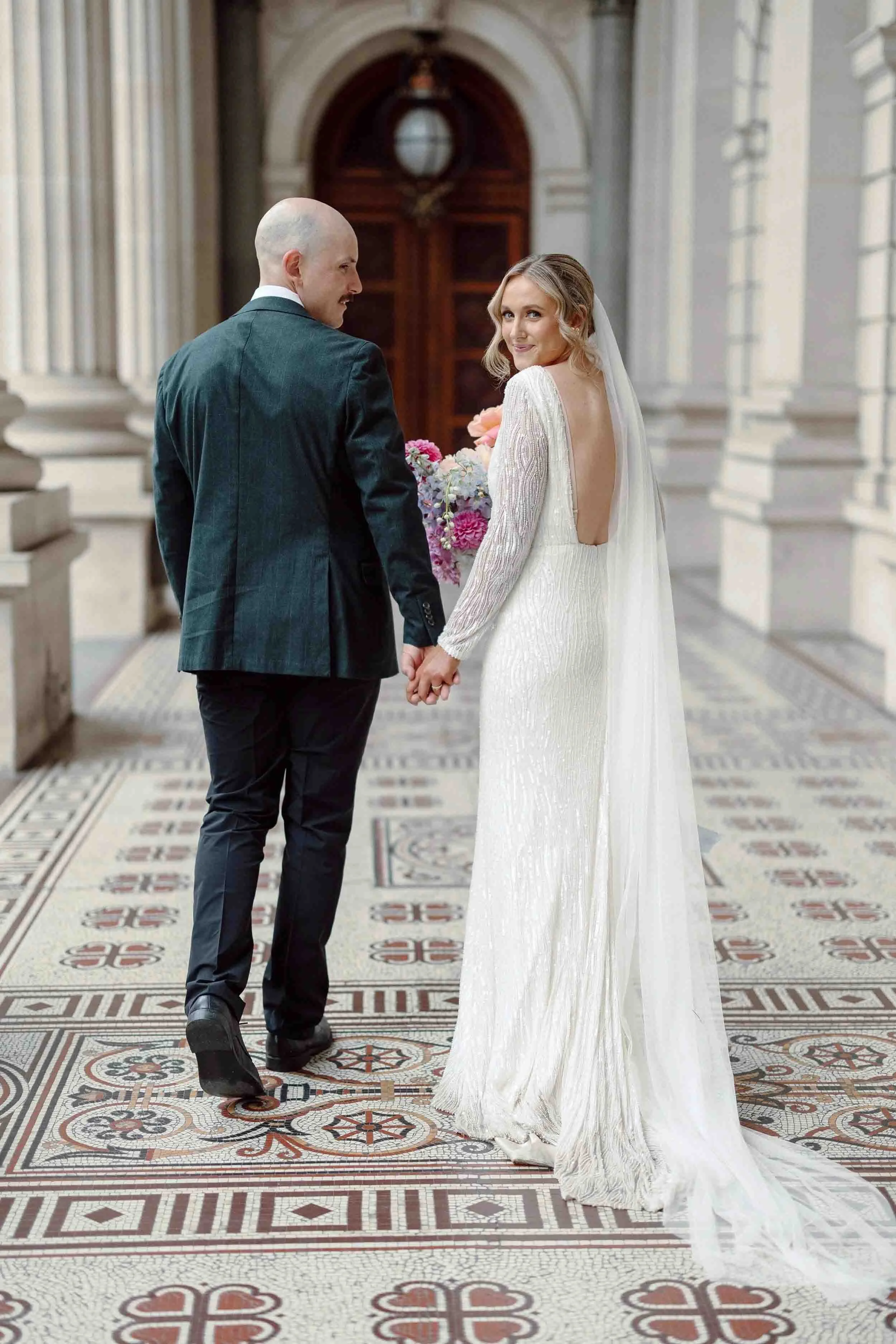 Aisling and Alex walk side by side at Fitzroy Gardens, her bouquet of dahlias, roses and orchids in full bloom.