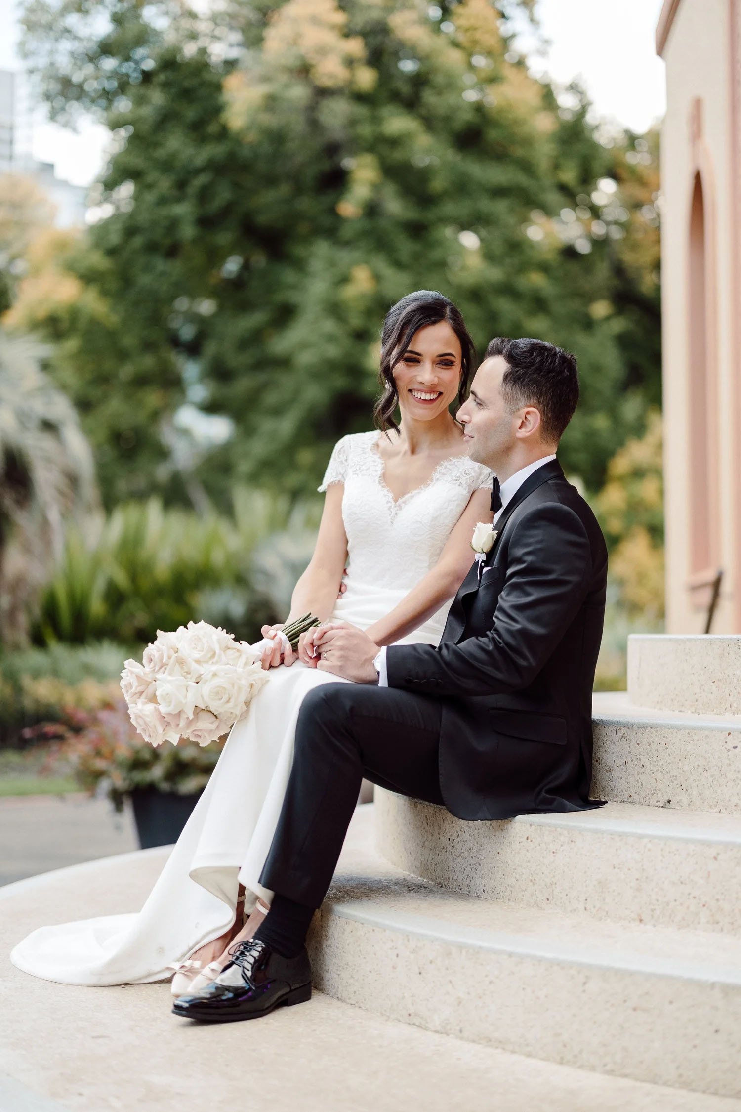 Kathryn laughs as Jason leans in on the stone steps at Fitzroy Gardens, autumn elms behind and white roses in her lap.