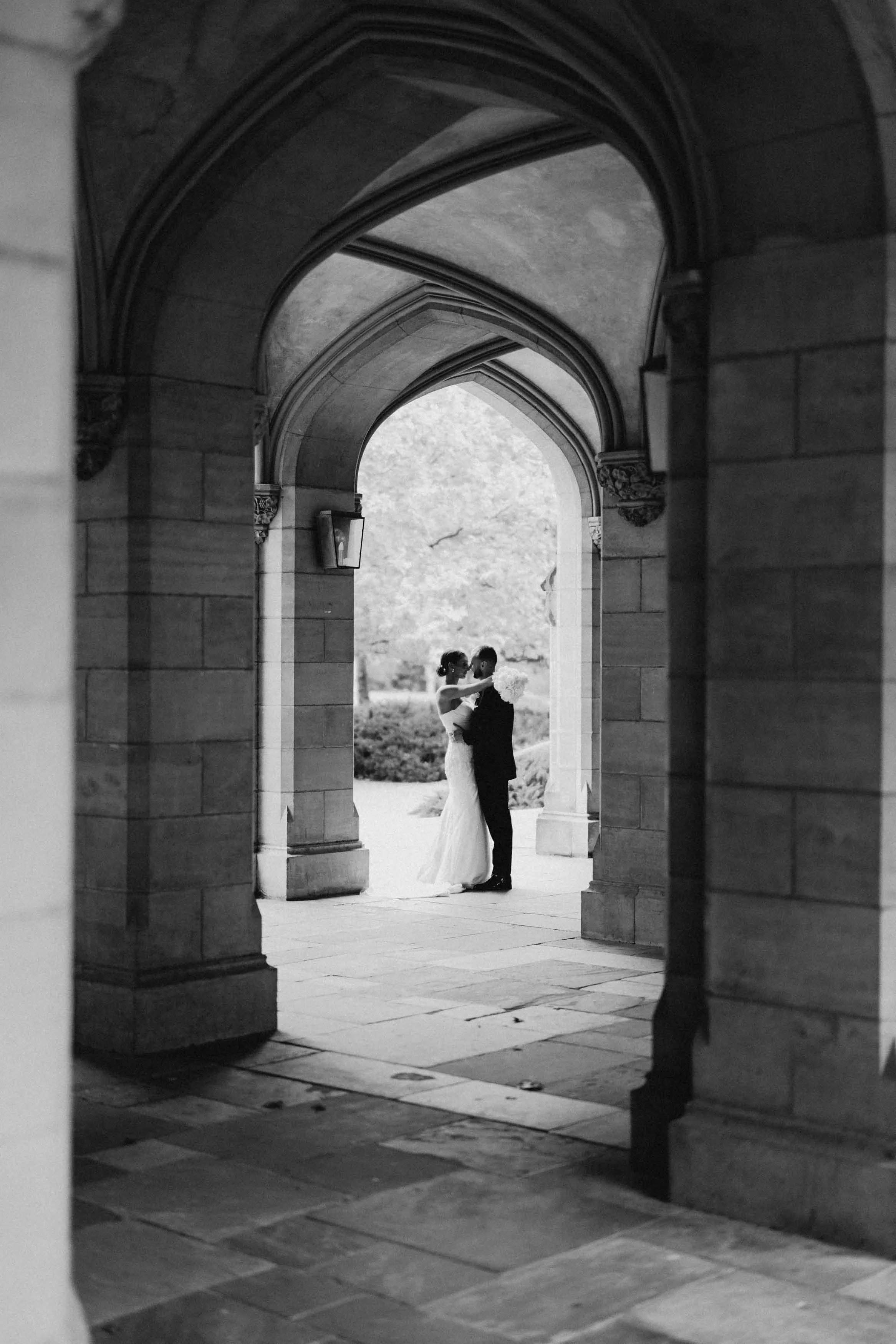 Framed by the gothic stone arches of the University of Melbourne cloisters, a bride and groom stand close together in the courtyard beyond.