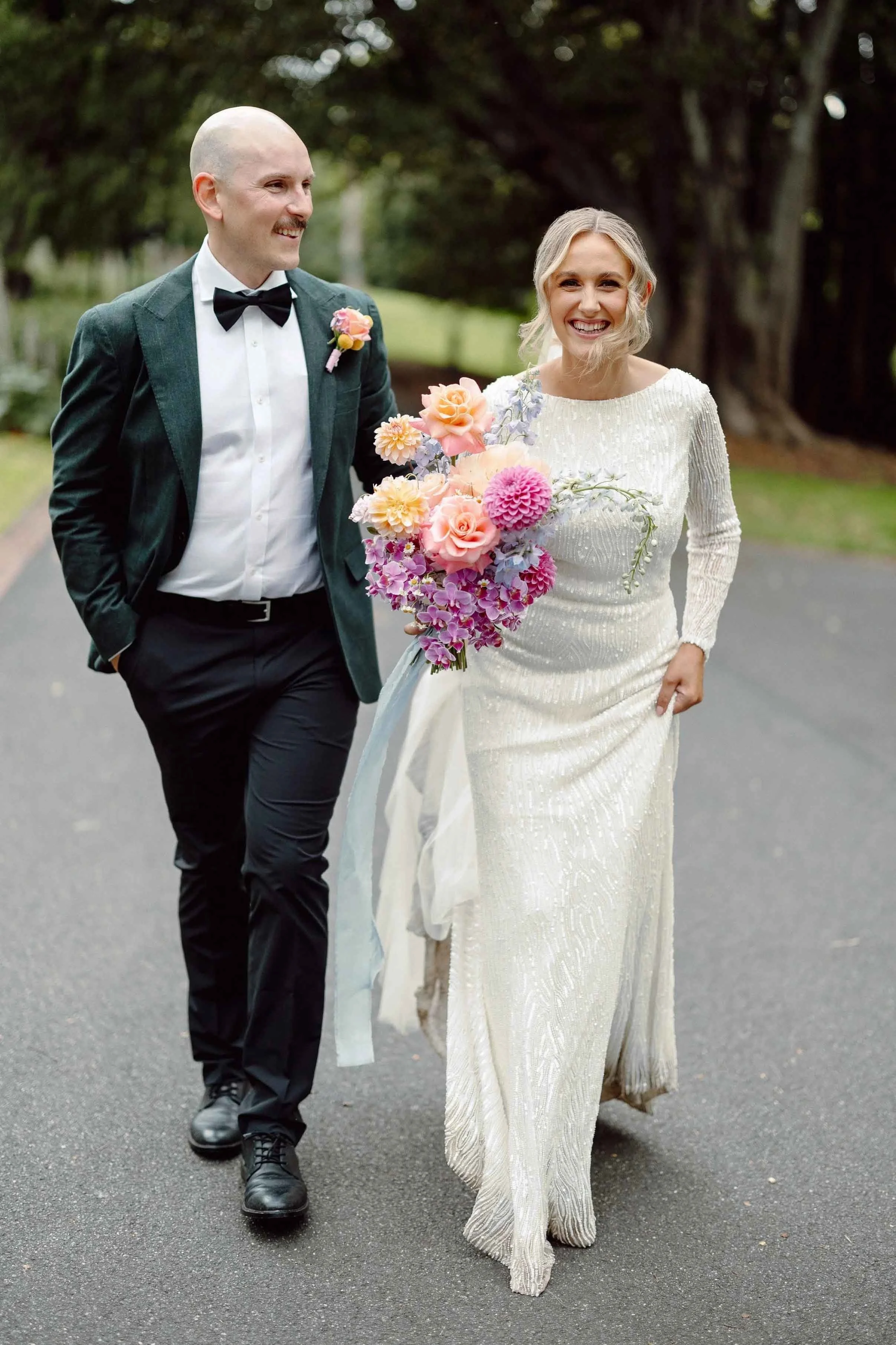 Aisling and Alex walk a tree-lined path at Fitzroy Gardens, her bouquet of dahlias and roses trailing blue ribbon.
