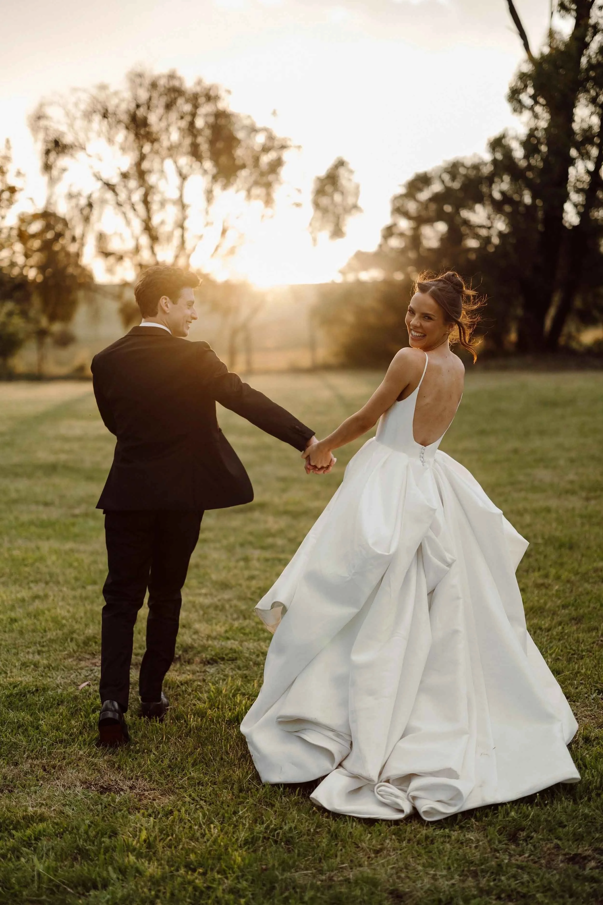 Ruby and Jacob run hand in hand through a gum-lined paddock at golden hour, her silk ballgown trailing behind her.
