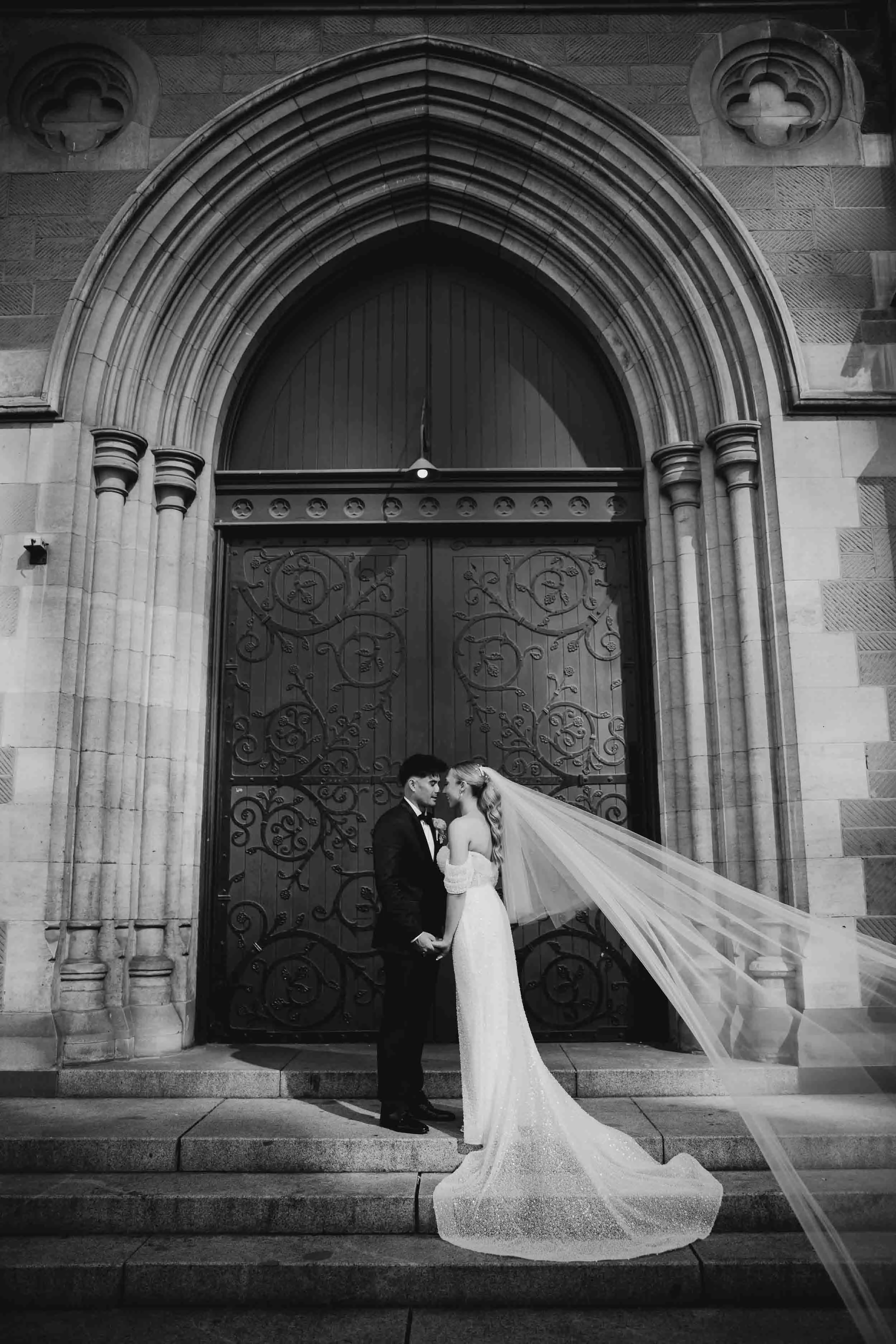 Laura's veil sweeps across the steps as she and Beau stand before the carved gothic doors of a bluestone cathedral in Melbourne's CBD.