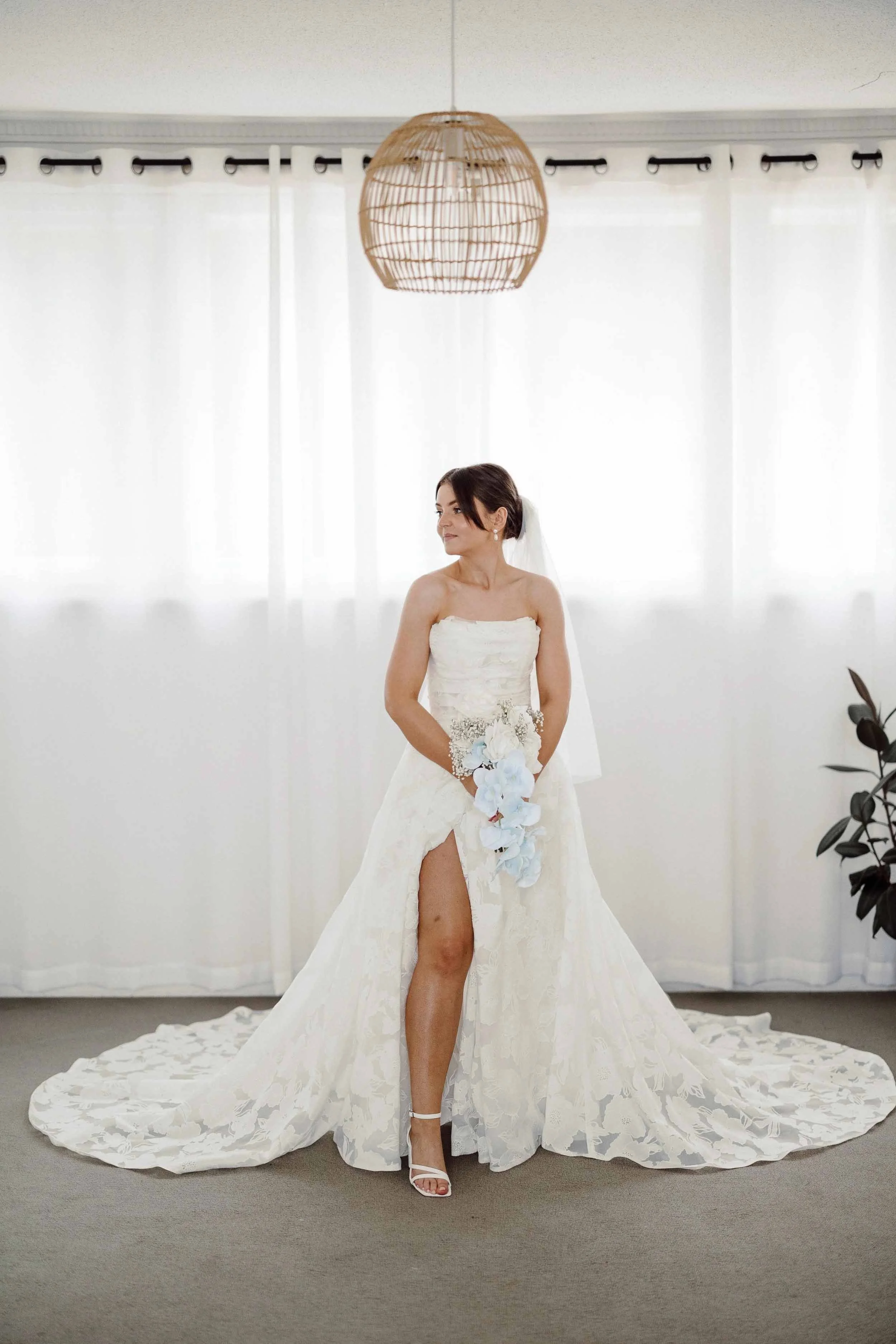 Amanda stands in the morning light before the ceremony, her lace gown spread across the floor and blue orchids in her hands.