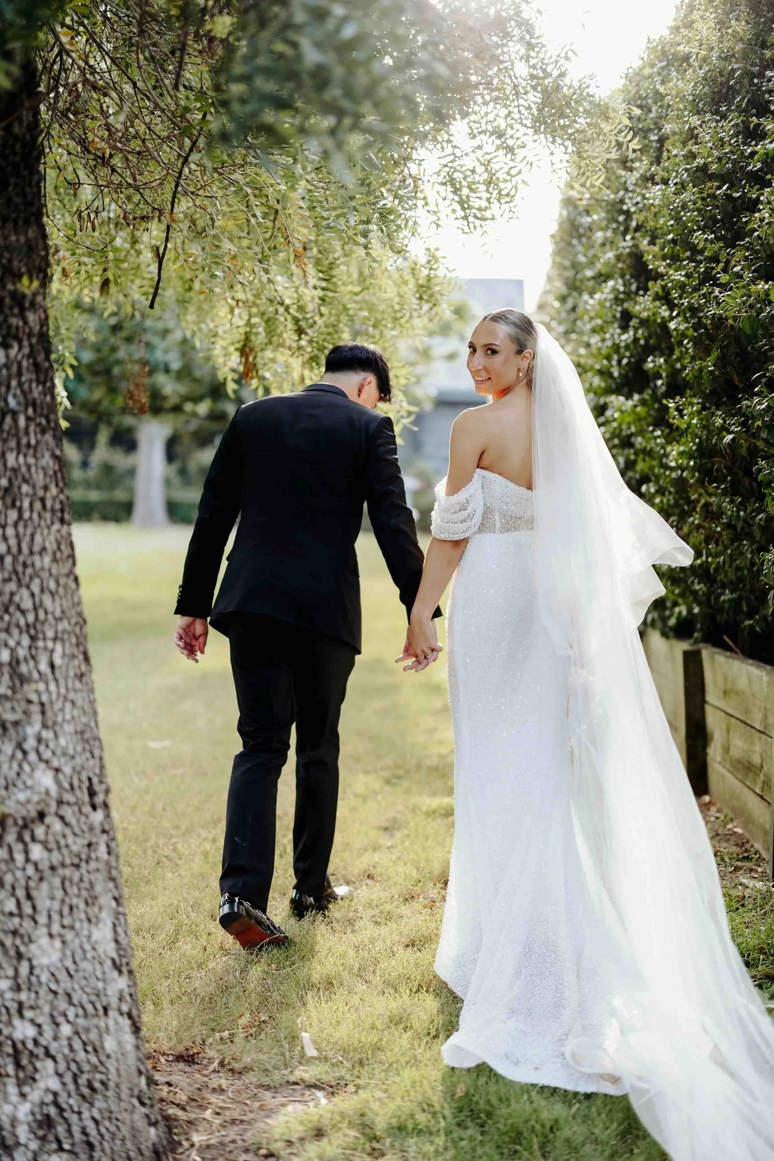 Laura and Beau walk together through the gardens at Meadowbank Estate, her veil catching the afternoon light.