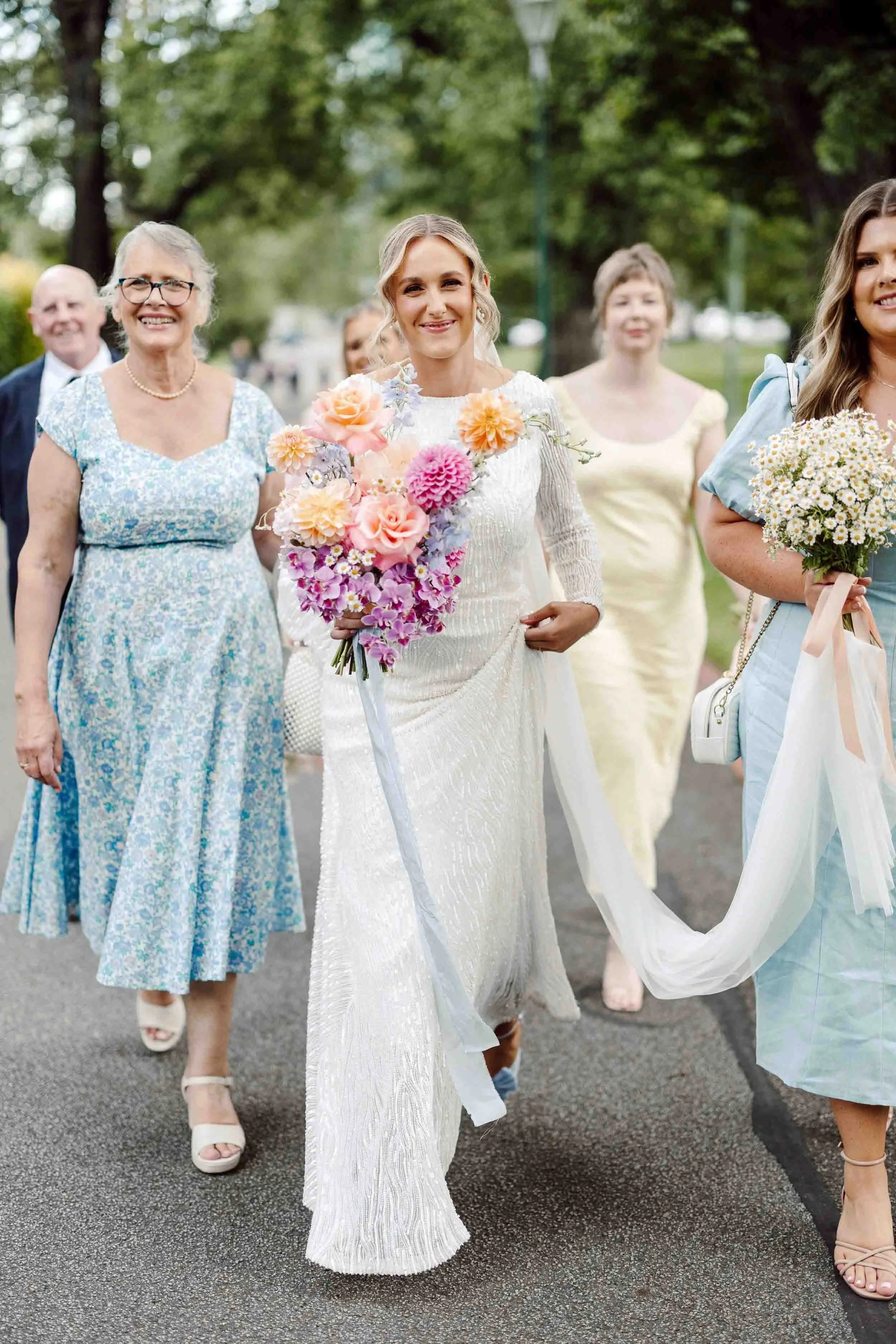 Aisling arrives for the ceremony at Fitzroy Gardens, smiling, her family and bridesmaids walking beside her with her veil gathered in their arms.