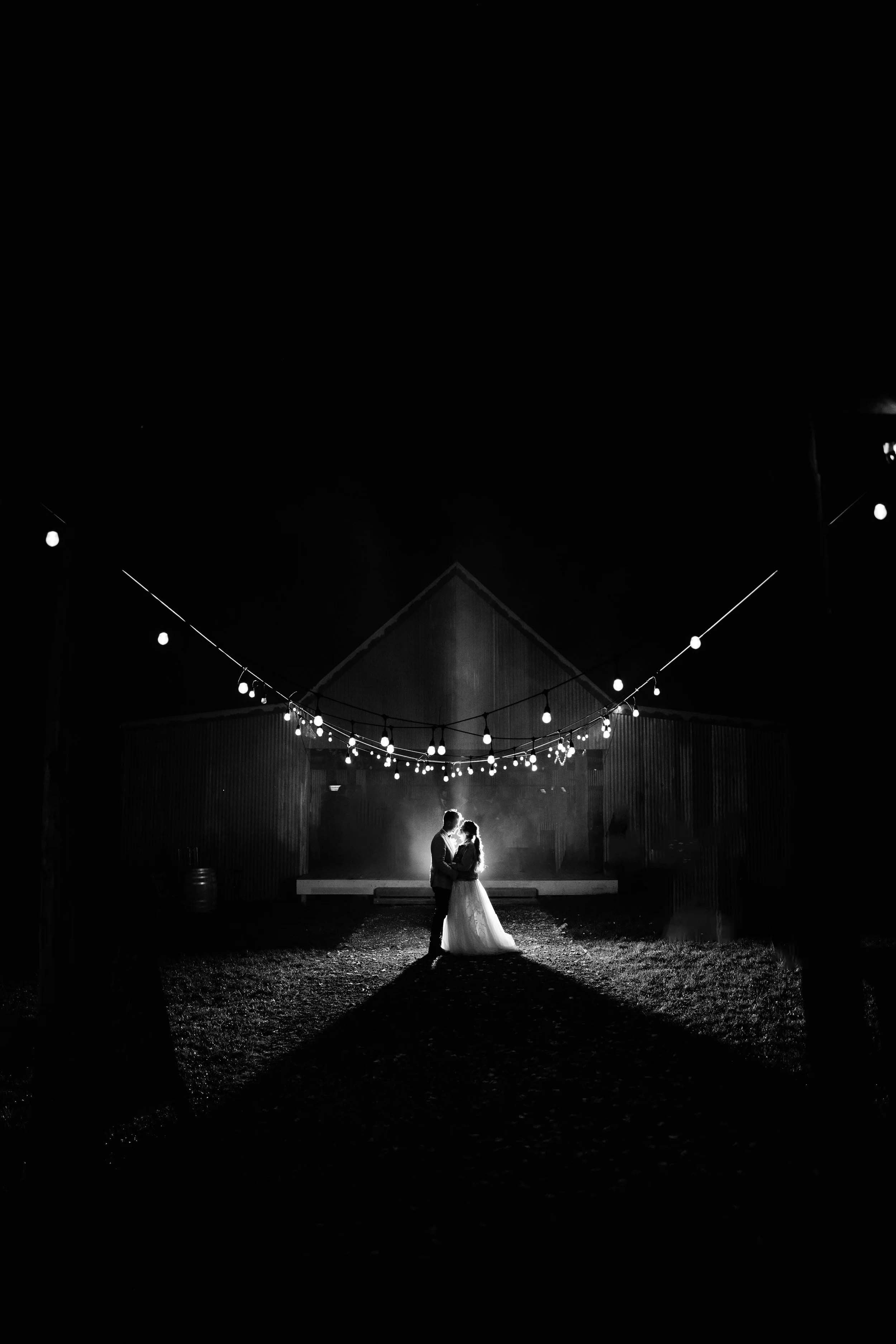 A bride and groom stand backlit beneath festoon lights outside a corrugated iron barn at night, their shadow stretching toward the camera.