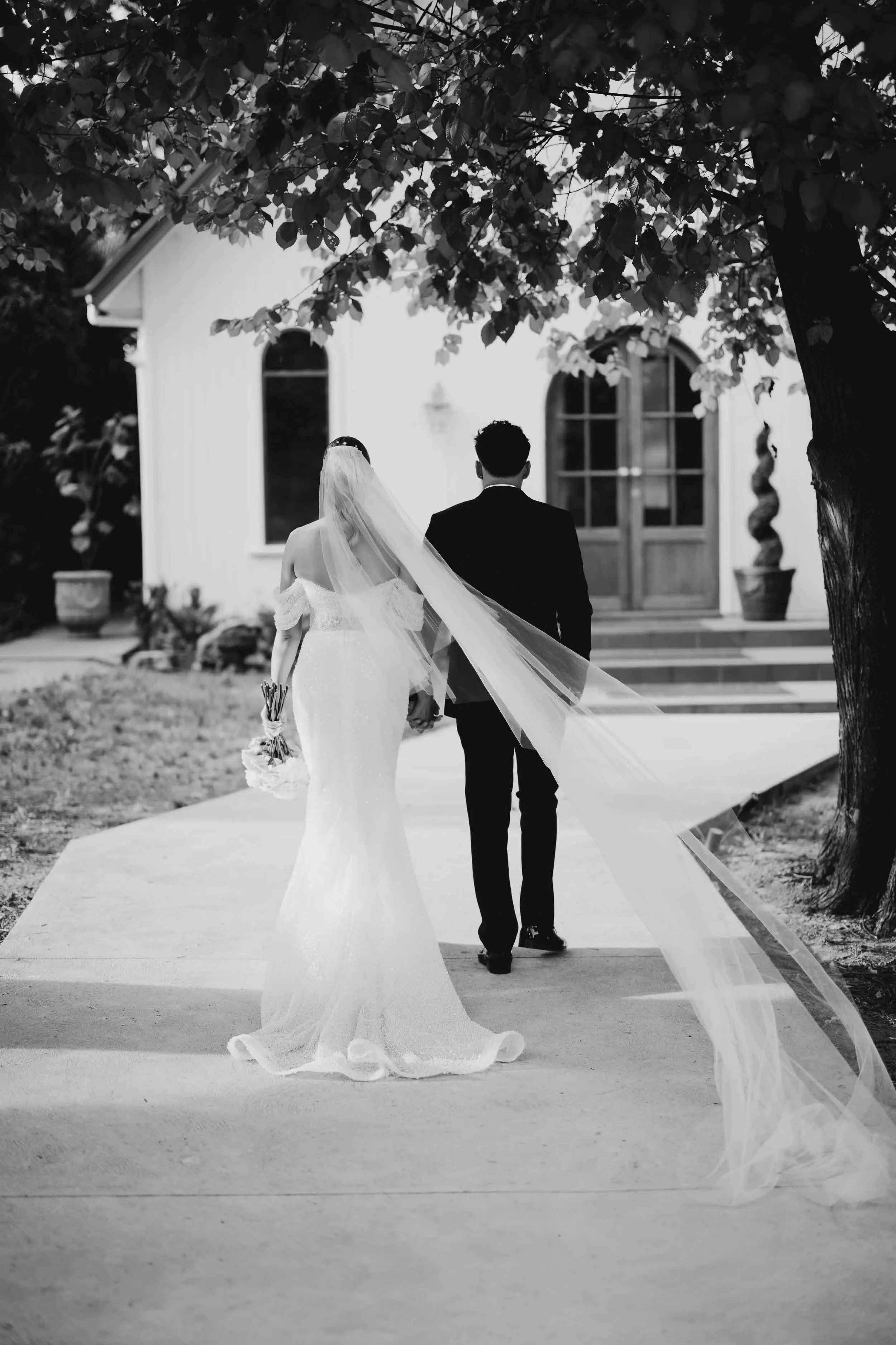 In black and white, Laura's veil streams behind her as she and Beau walk hand in hand toward the arched doors of the chapel at Meadowbank Estate.