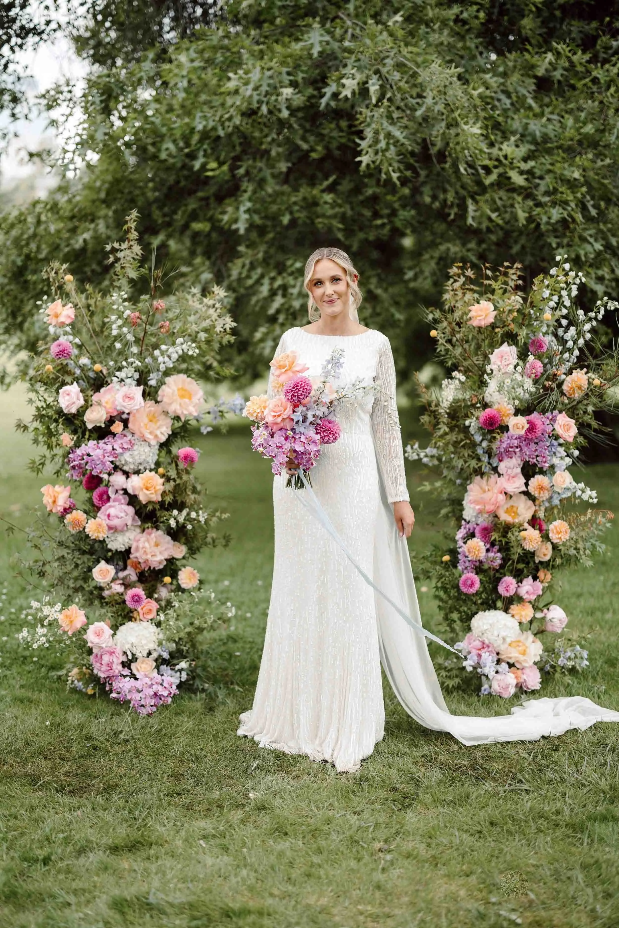 Aisling stands between the two floral installations at Fitzroy Gardens, her beaded gown and veil trailing across the grass.