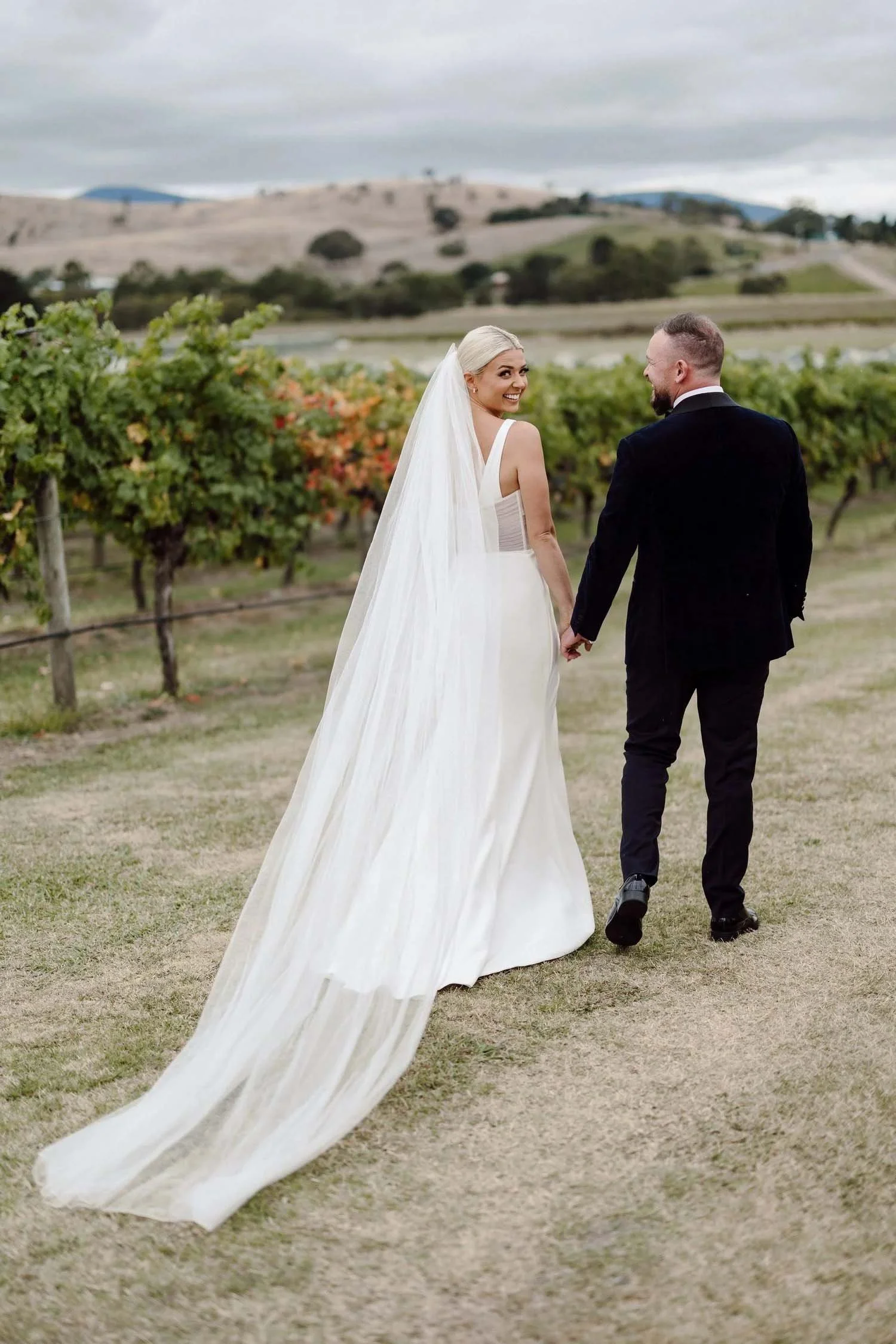 Maddison glances back over her shoulder as she and Michael walk the vines at Acacia Ridge, her cathedral veil trailing behind.