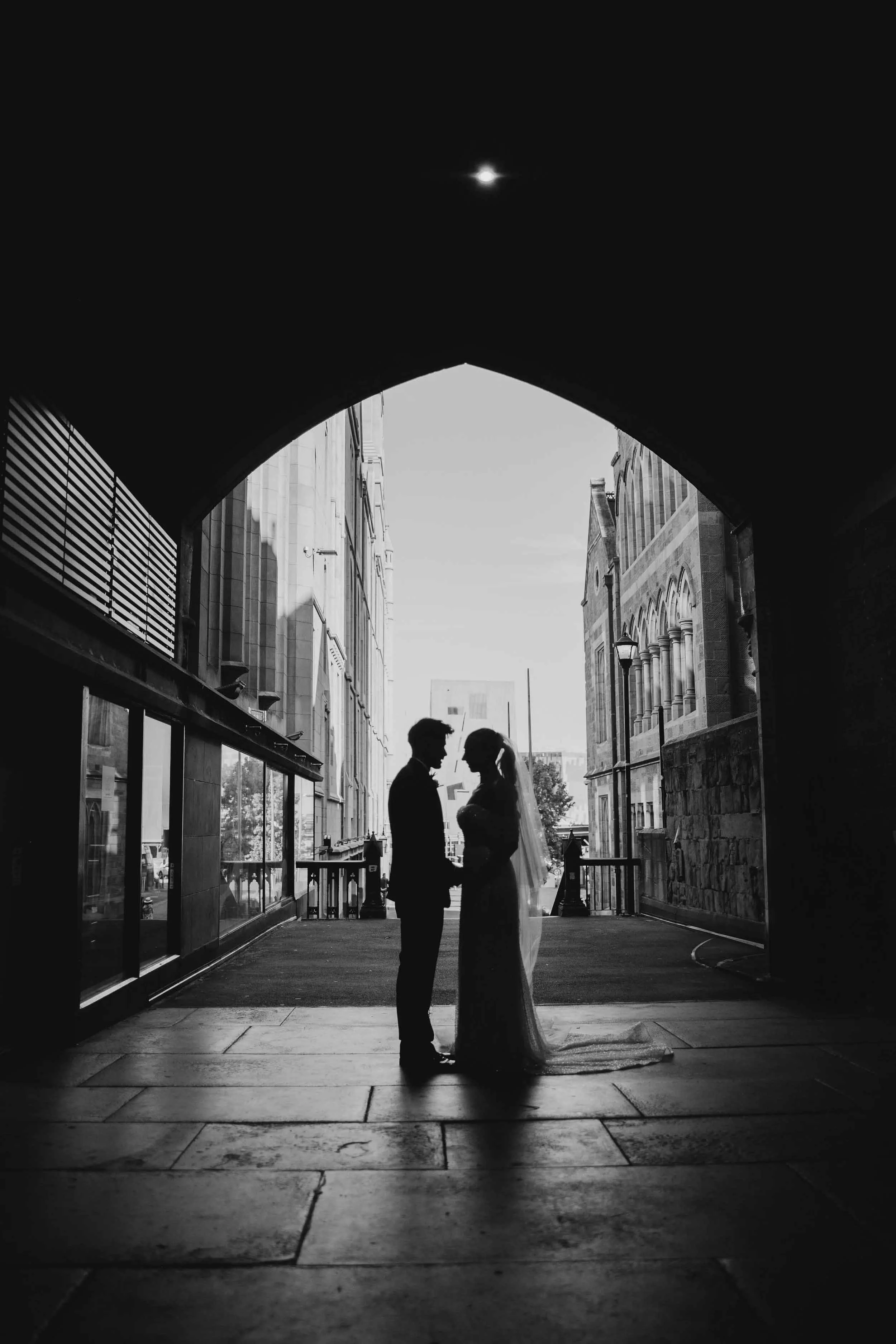 Laura and Beau stand hand in hand in silhouette beneath a stone archway, the bluestone buildings of Melbourne's CBD framed beyond them.