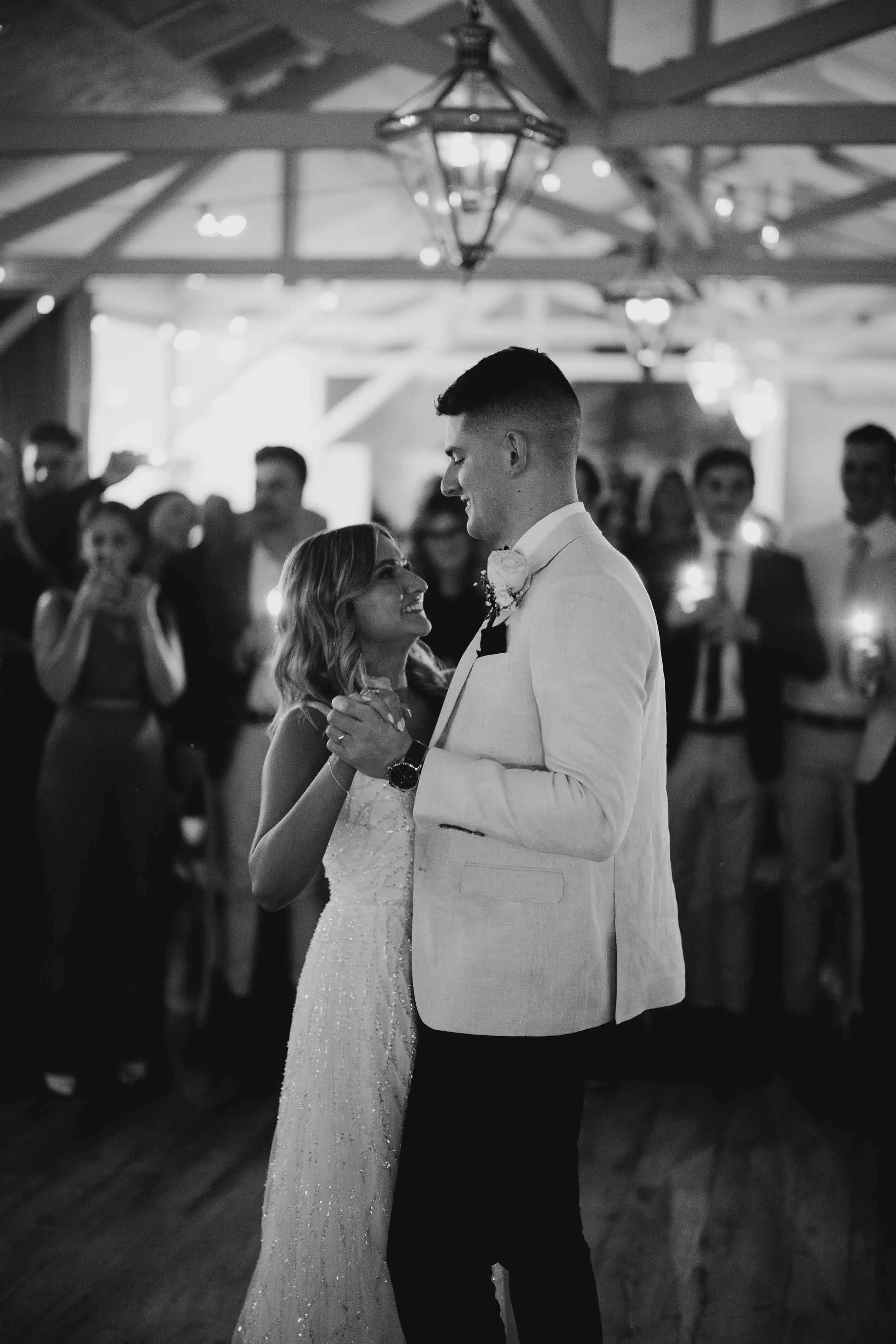 Alt text:

Nicola looks up at Nick during their first dance at Bramleigh Estate, guests circled around them holding sparklers beneath the timber trusses.