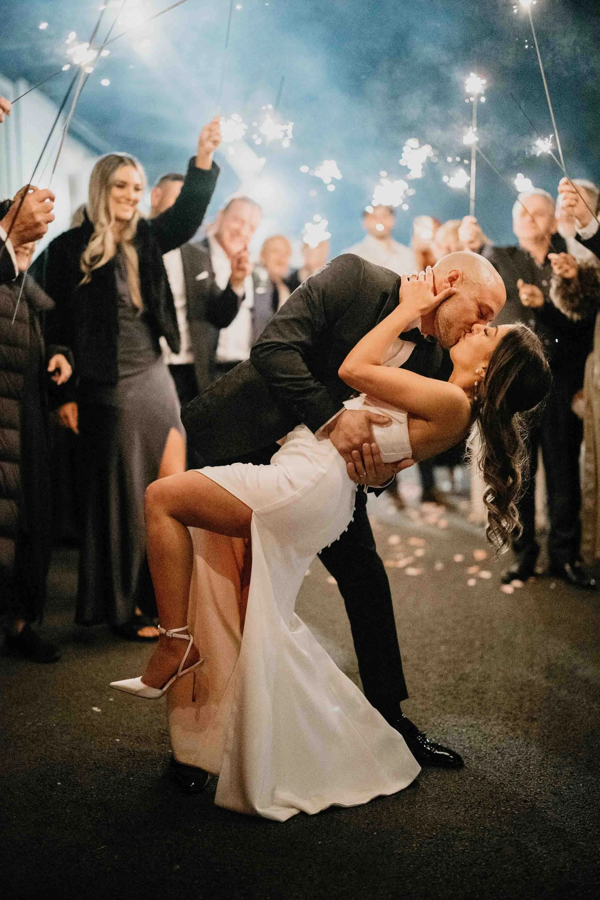 Dean dips Rachel into a kiss beneath an arch of sparklers held by their guests at Bramleigh Estate.