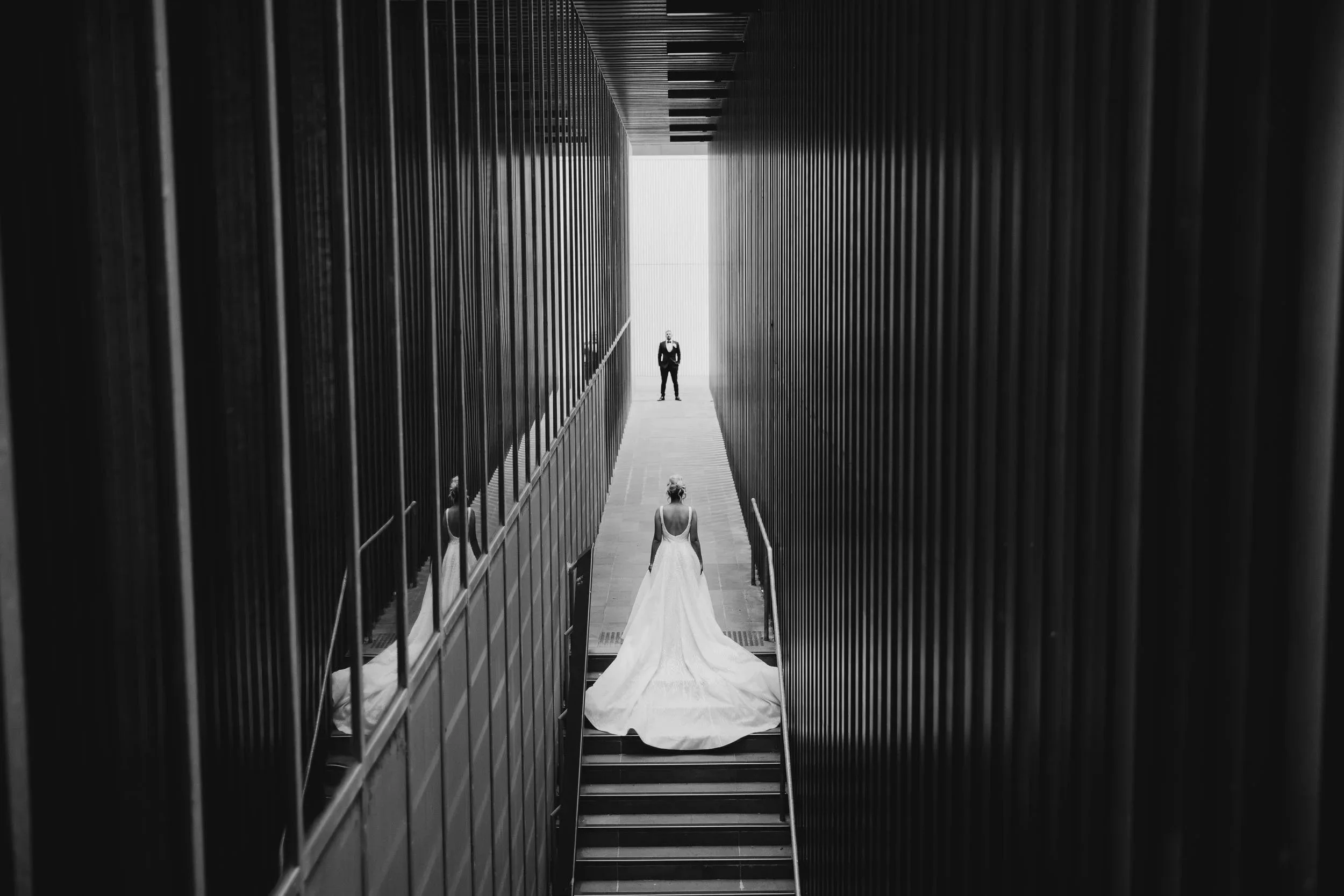 In black and white, a bride pauses on the stairs in a corridor of dark vertical fins at RMIT University, her groom waiting at the far end.