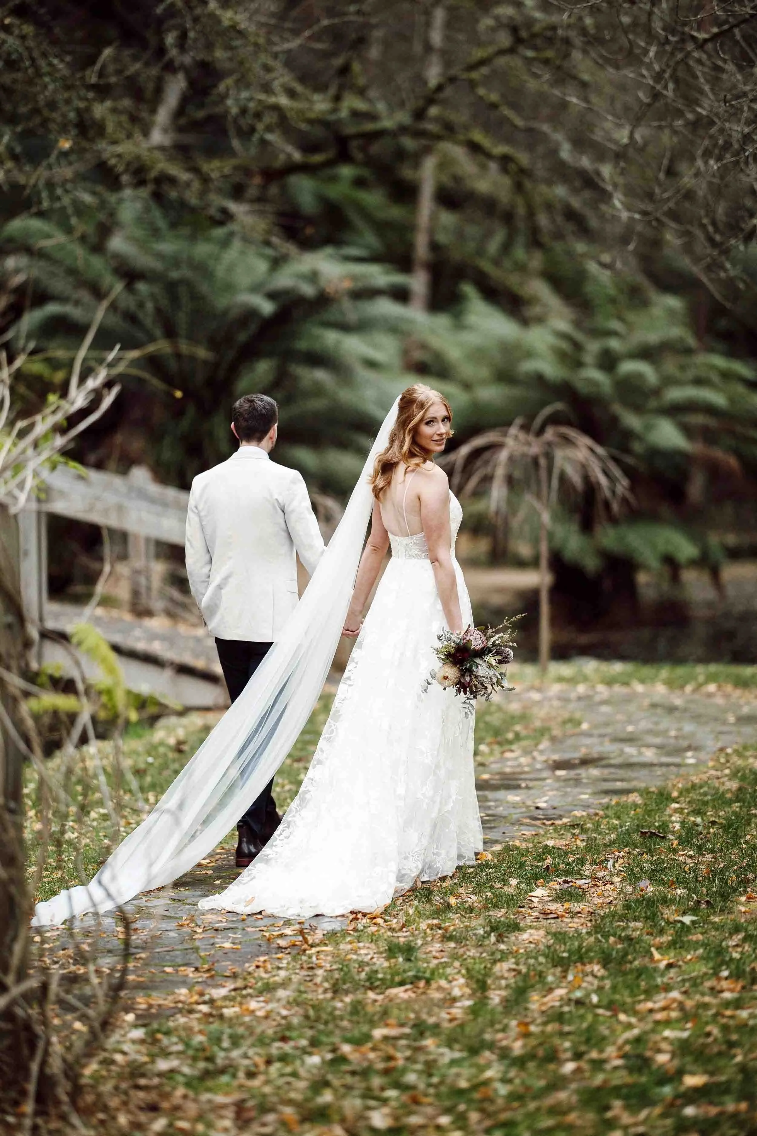 Tayla looks back over her shoulder as she and Scott walk the stone path at Alfred Nicholas Memorial Gardens in autumn, fallen leaves and tree ferns around them.
