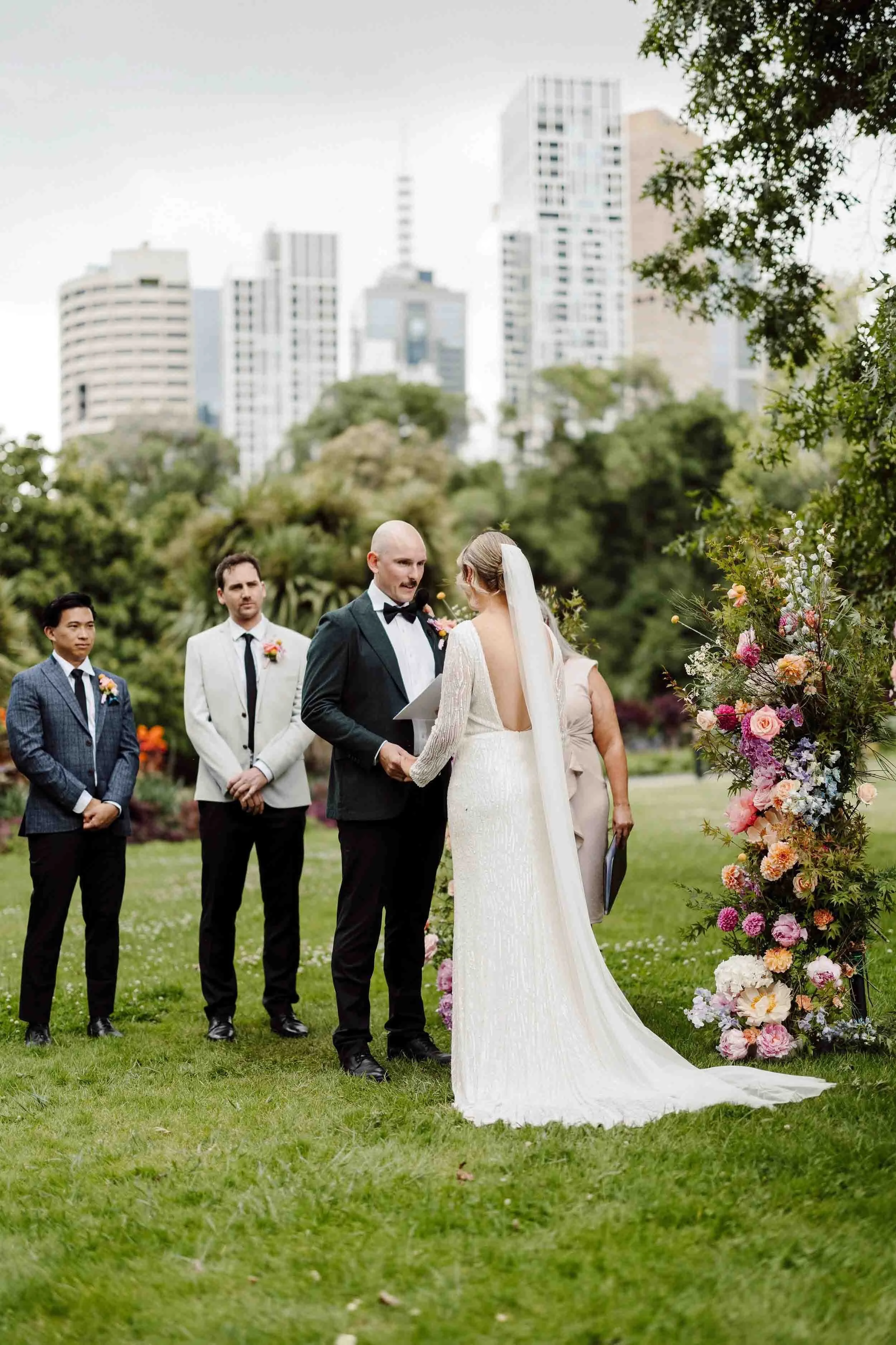 Aisling and Alex exchange vows beside a floral installation at Fitzroy Gardens, the Melbourne city skyline rising behind the trees.
