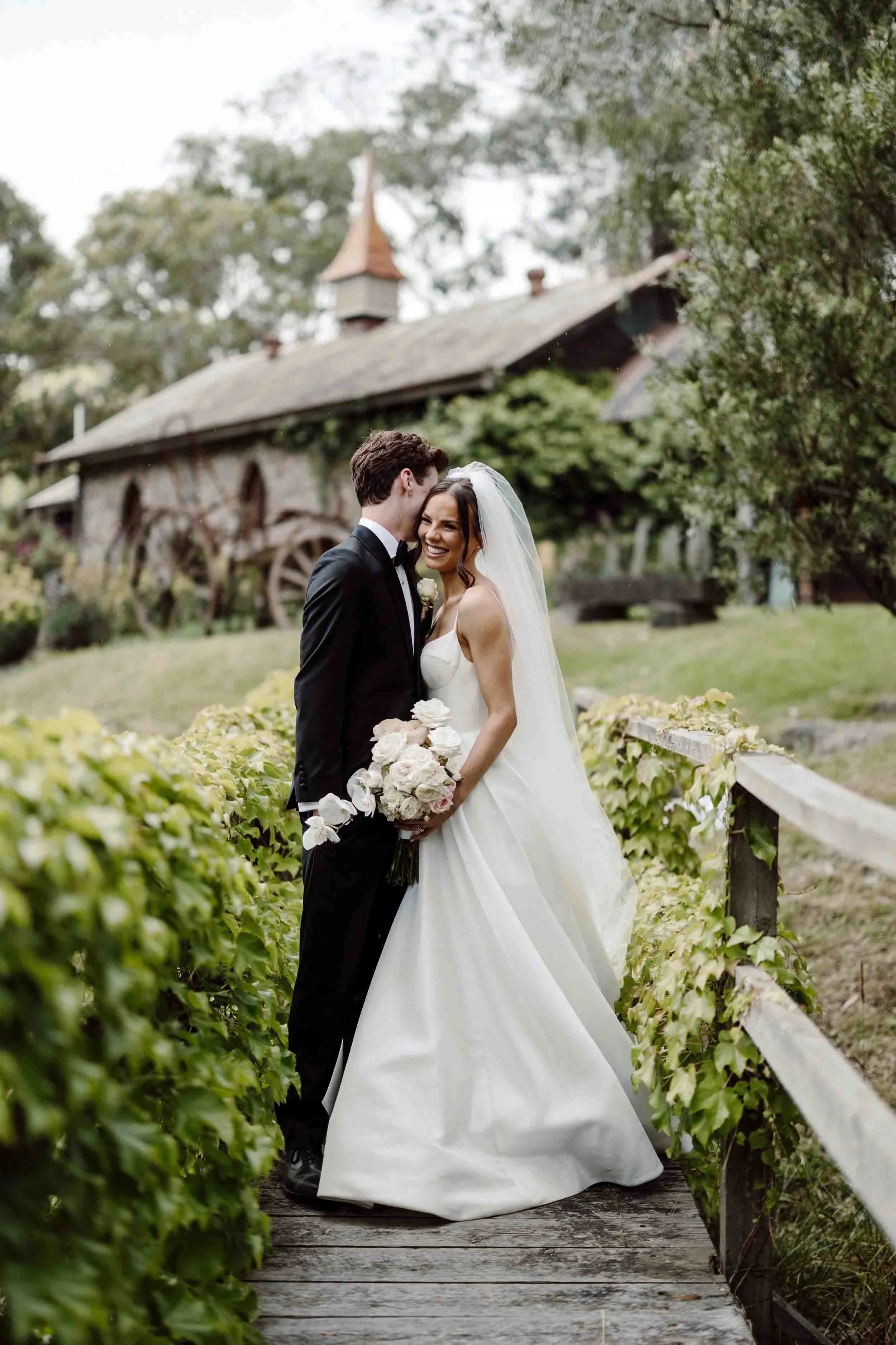Ruby leans into Jacob on the ivy-lined timber bridge at Inglewood Estate, the stone barn and its weathervane behind them.