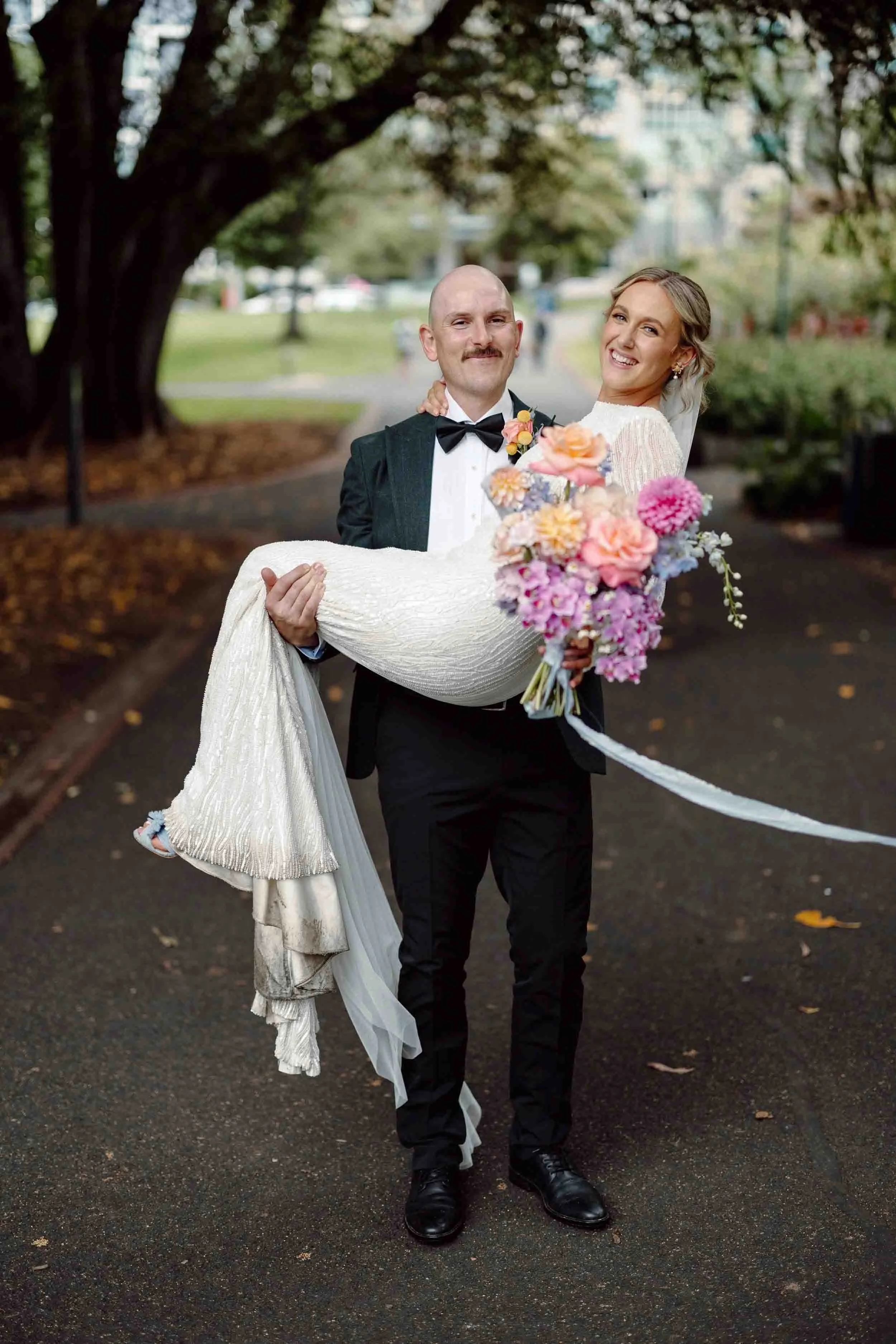 Alex carries Aisling along a path at Fitzroy Gardens, both of them laughing, her bouquet ribbon streaming and autumn leaves on the ground.