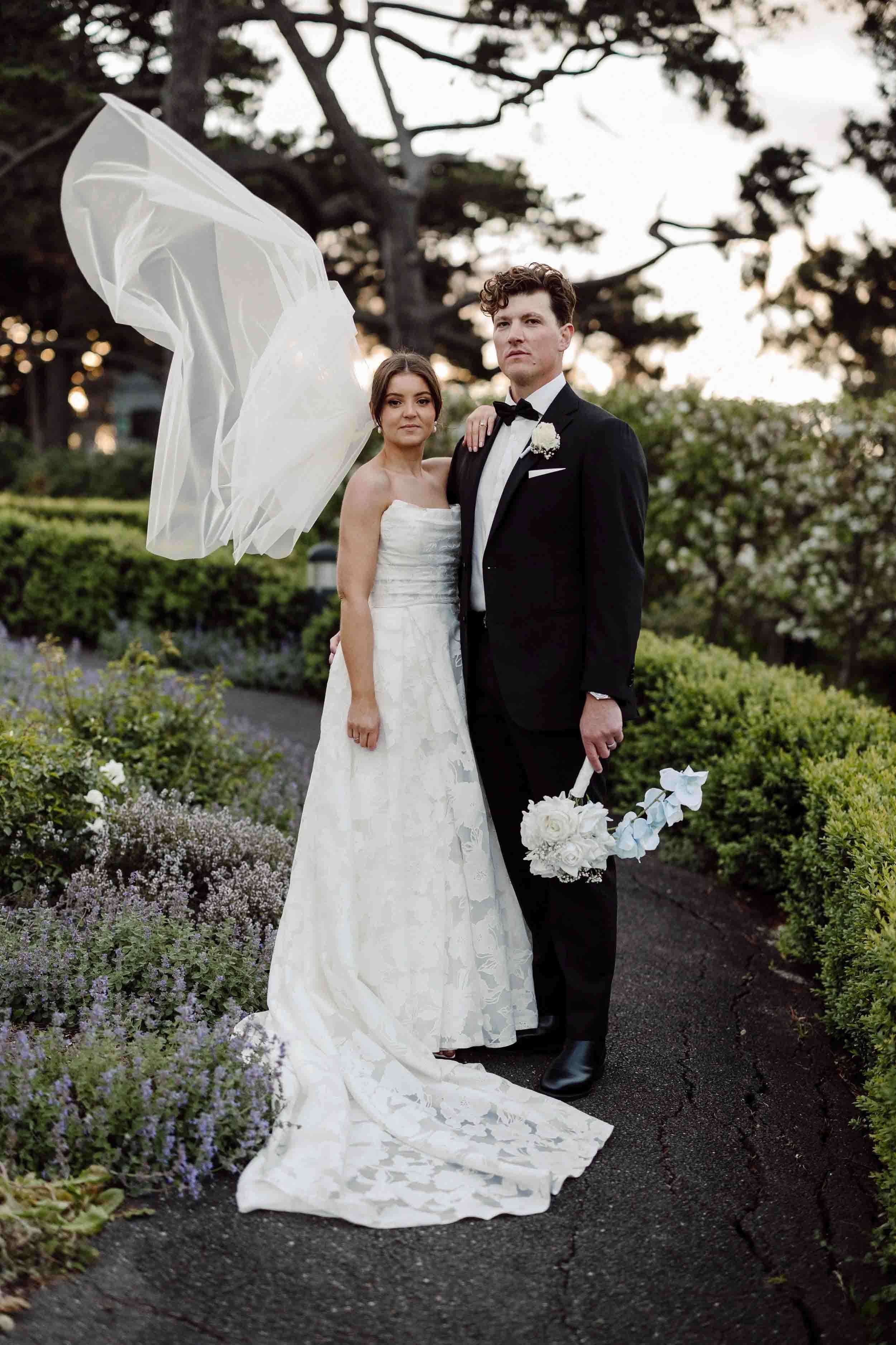 Amanda's veil lifts on the wind as she and Jesse stand together on the garden path at Farm Vigano, lavender and clipped hedges around them.