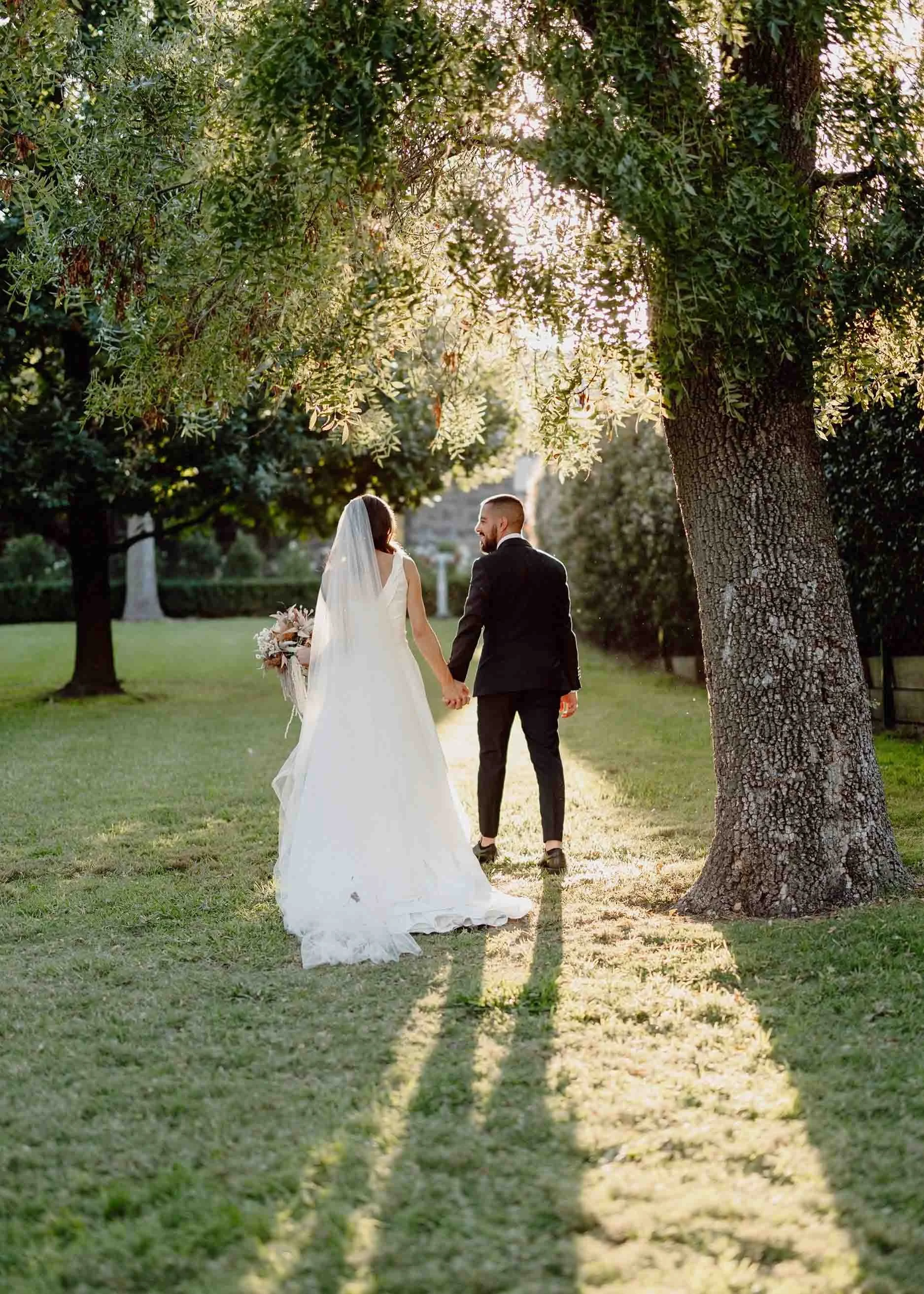 Julia and Luca walk hand in hand beneath the olive trees at Meadowbank Estate, long shadows stretching across the grass in the late light.