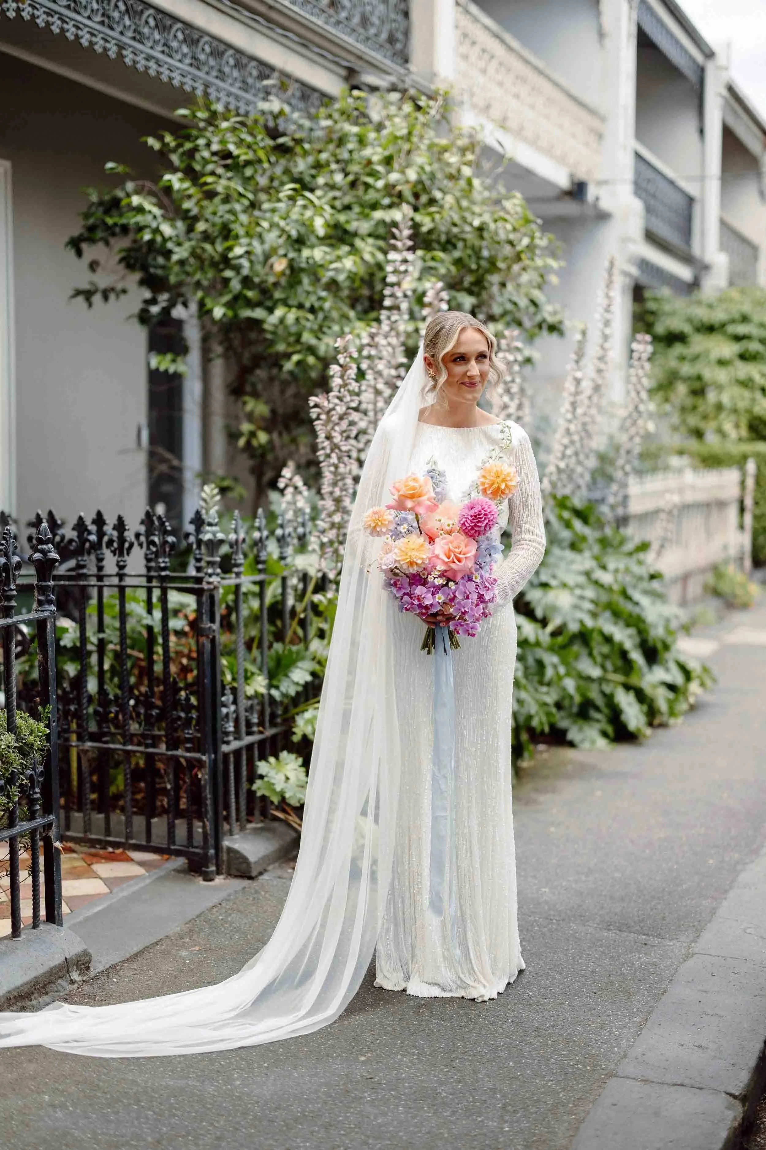 Aisling stands on the footpath outside an East Melbourne terrace, her cathedral veil pooling behind her and a bouquet of dahlias and roses in her arms.