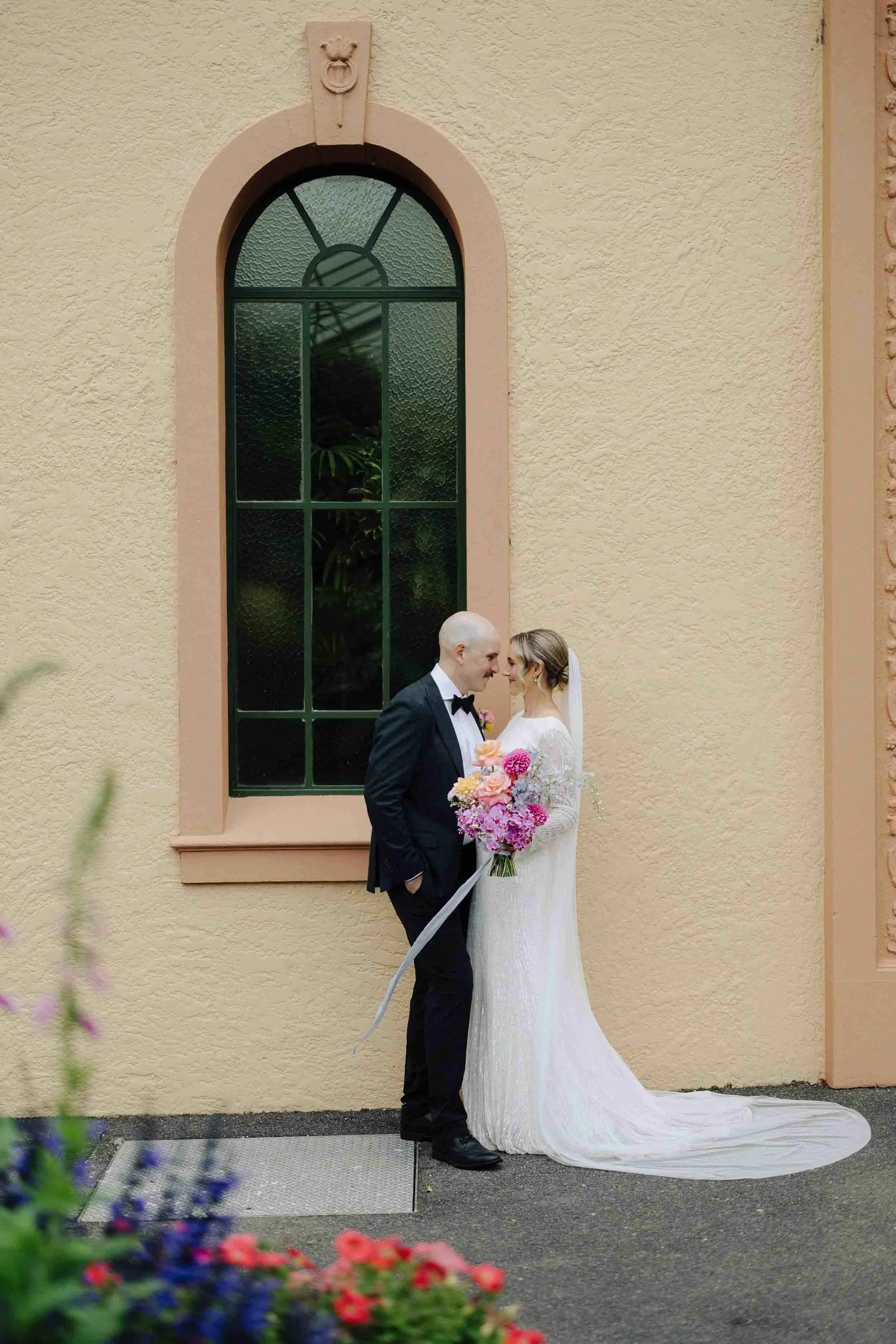 Aisling and Alex stand forehead to forehead beside the arched green window of the Conservatory at Fitzroy Gardens, garden beds in bloom around them.
