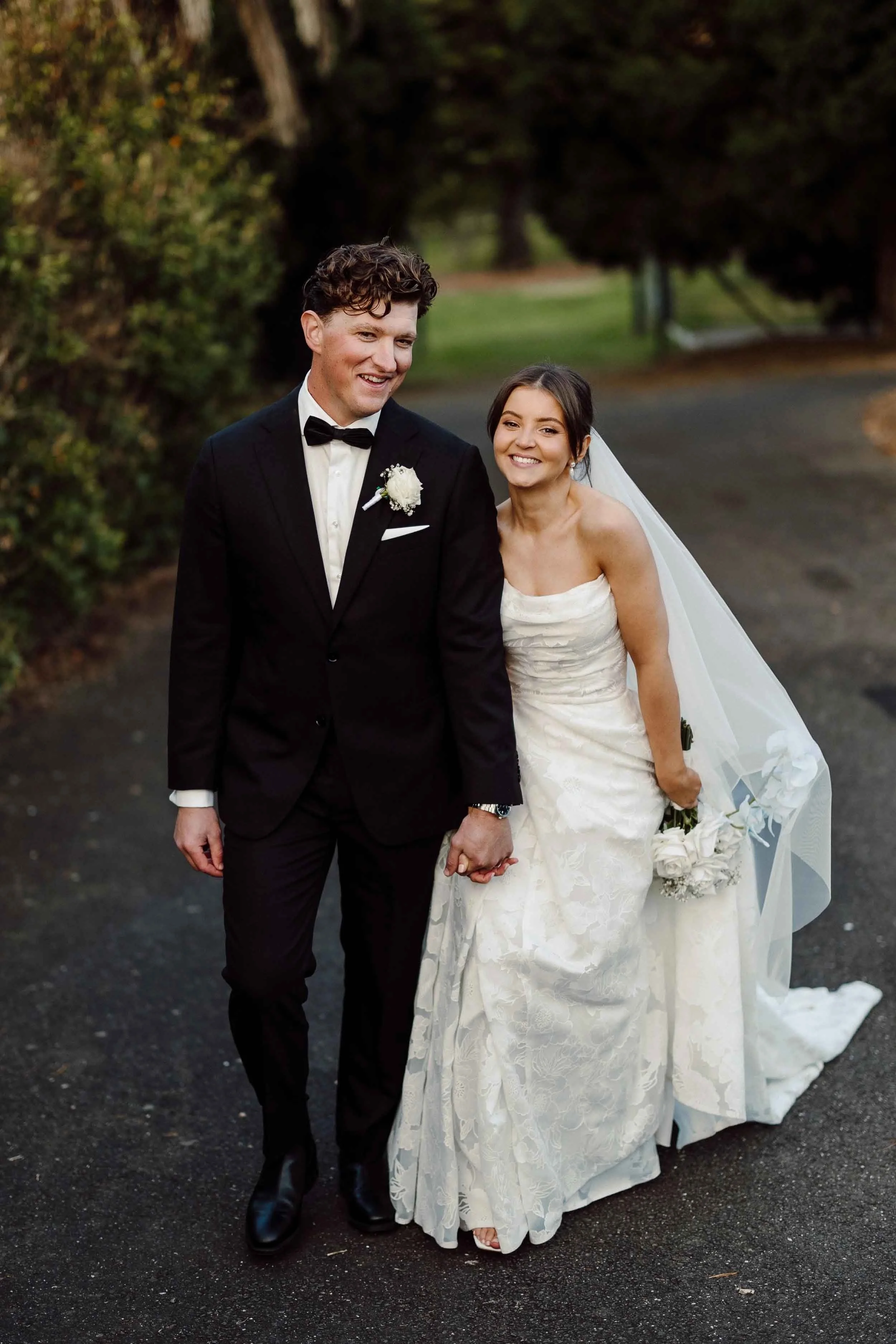 Amanda and Jesse walk hand in hand along the driveway at Farm Vigano, her lace gown and veil trailing beside her.