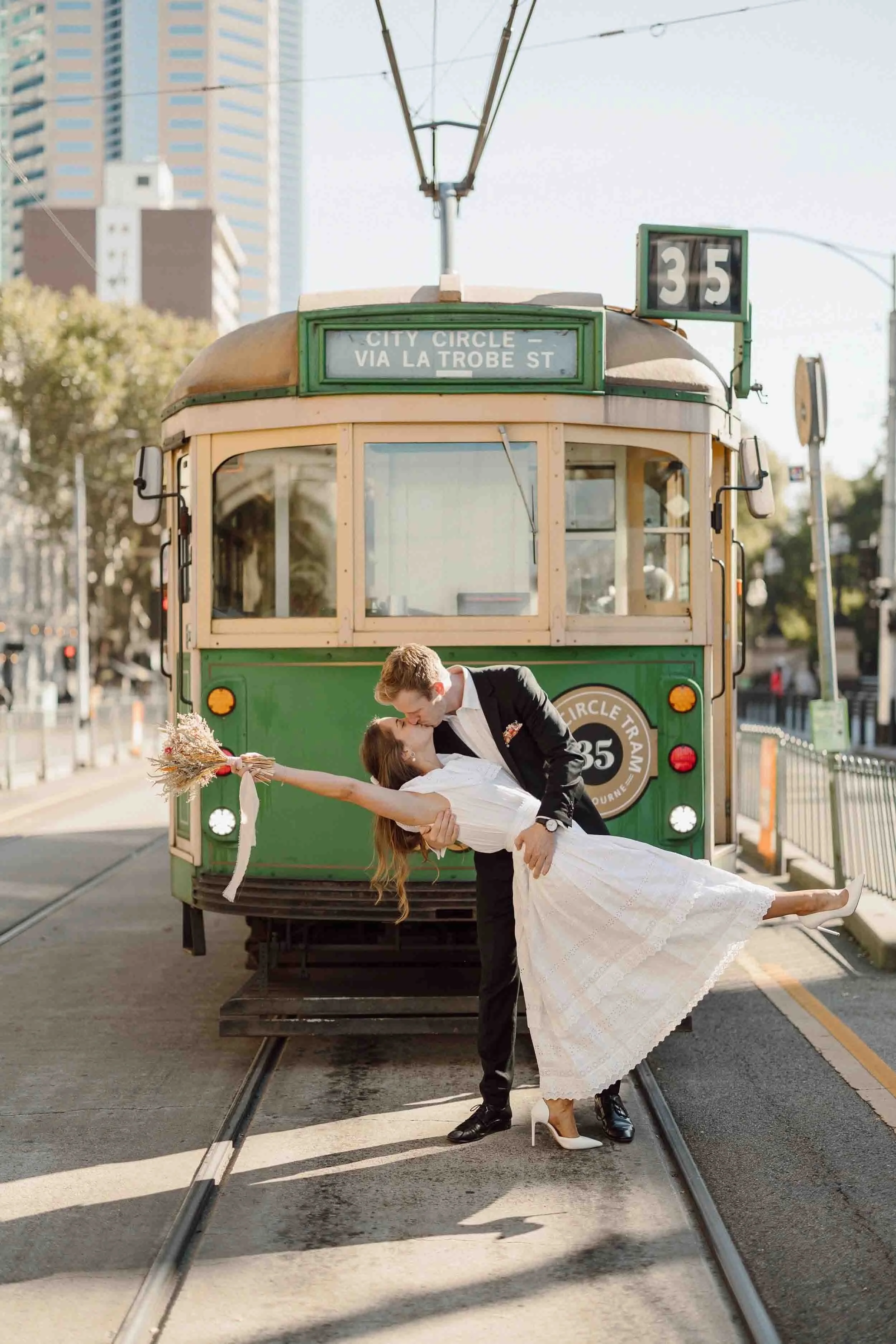 Tim dips Heloise into a kiss in front of the green City Circle tram out the front of parliament steps in Melbourne her dried bouquet out to the side.
