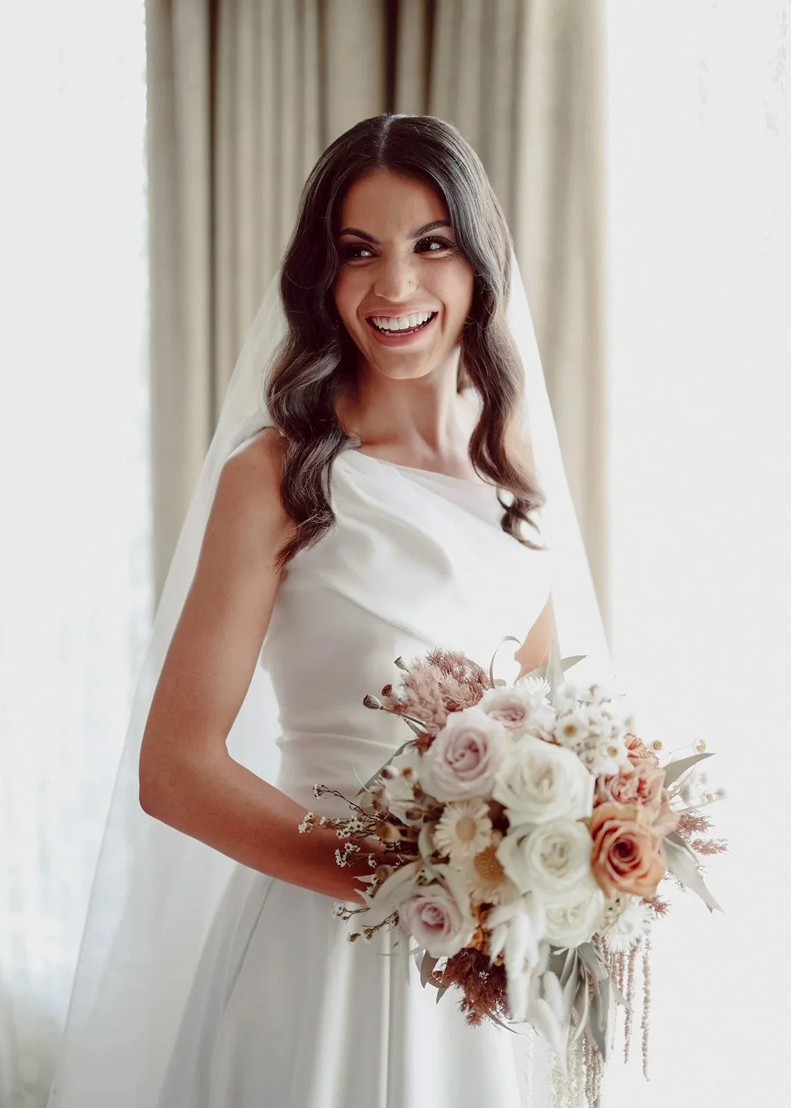 Julia laughs by the window in the morning light at Meadowbank Estate, her cowl-neck gown catching the light and a bouquet of blush roses and dried natives in her hands.
