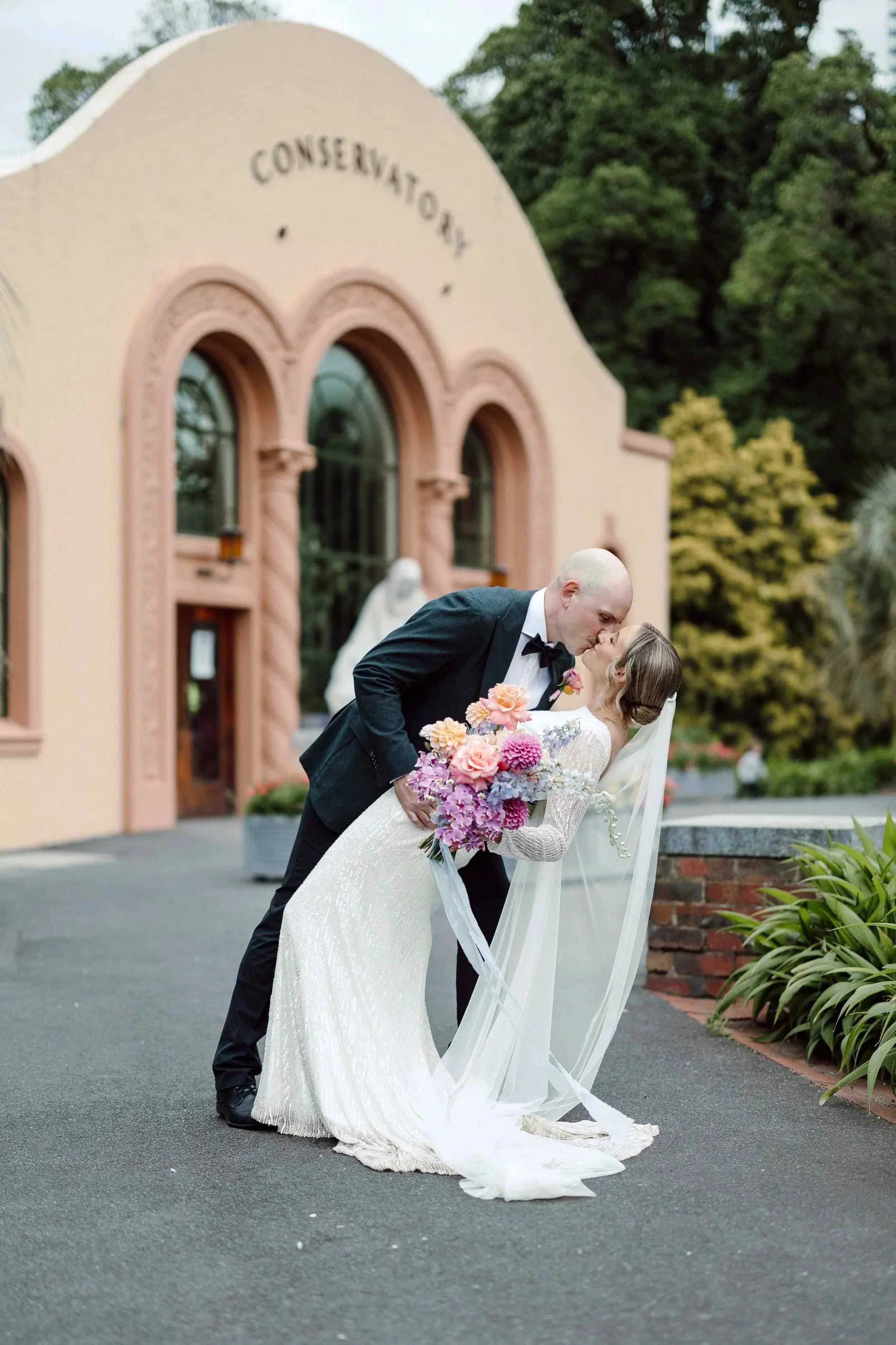 Alex dips Aisling into a kiss outside the Conservatory at Fitzroy Gardens, her veil sweeping the path and her bouquet held out beside them.