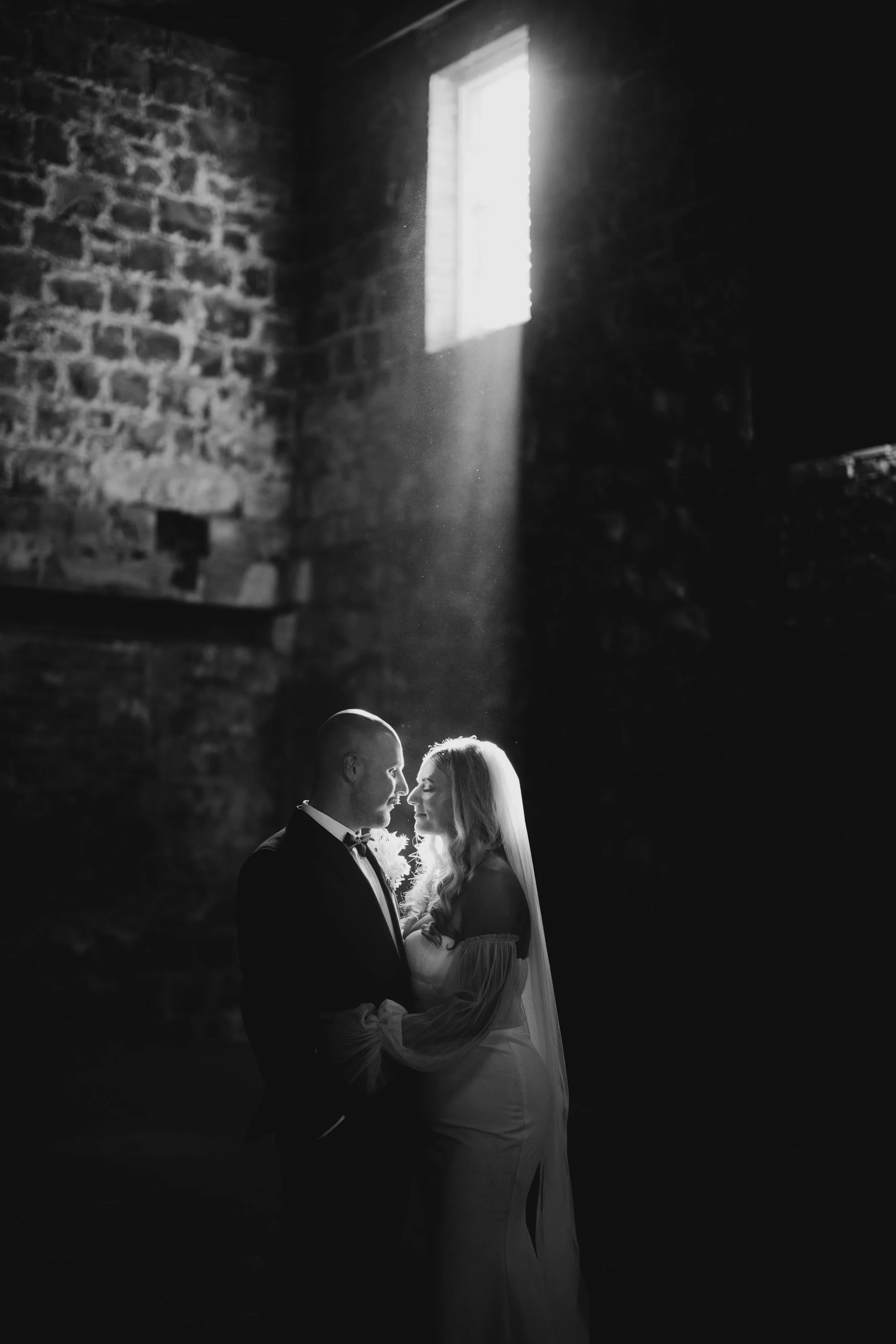 A single shaft of light from a high window falls across a bride and groom standing close together inside the stone barn at Providence Wines.